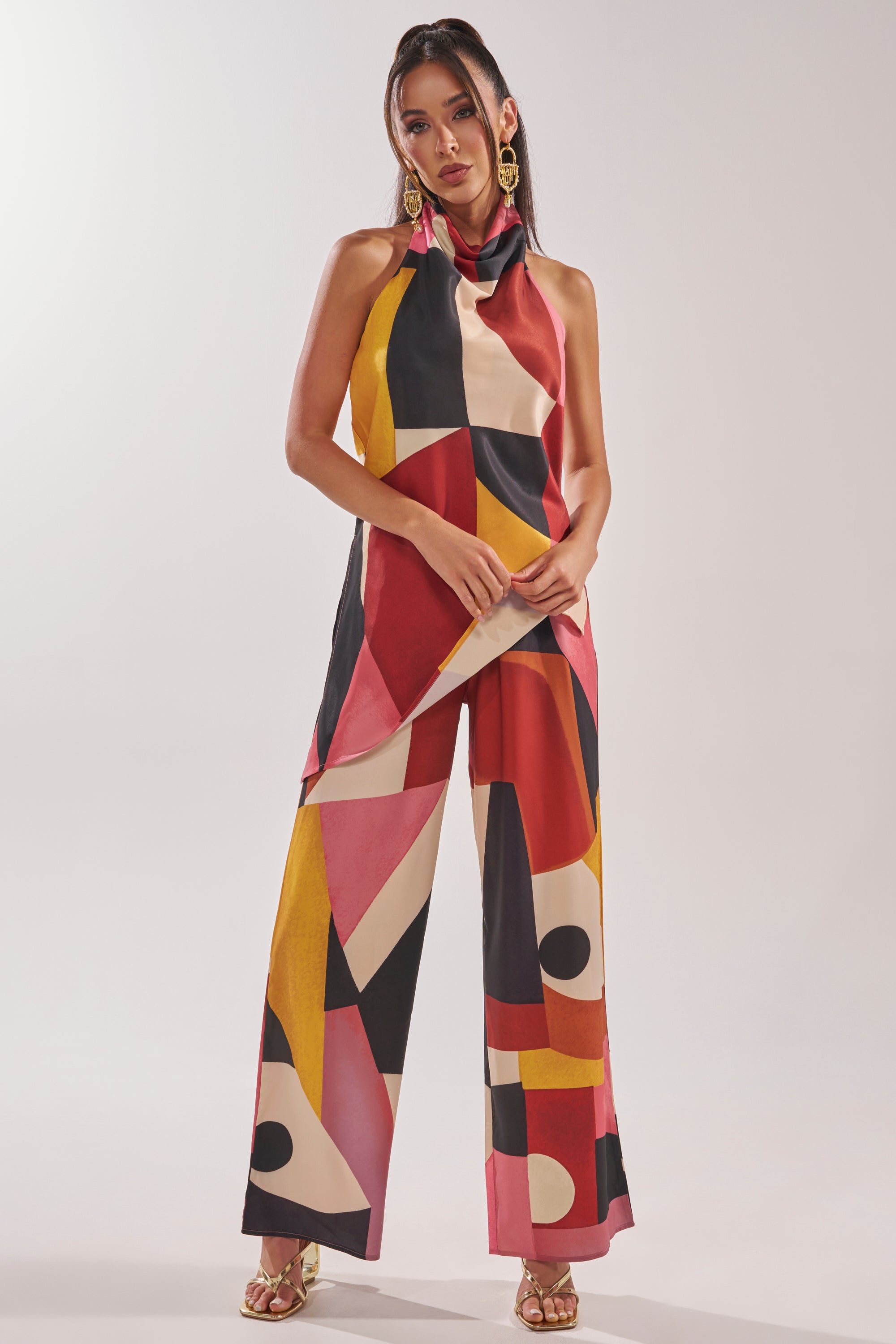 A woman wears the MIXED EMOTIONS TROUSER featuring a vibrant red, yellow, black, and white geometric print with a sleeveless, high-neck top, large earrings, and gold sandals—ideal vacation trousers for a chic getaway look. 6