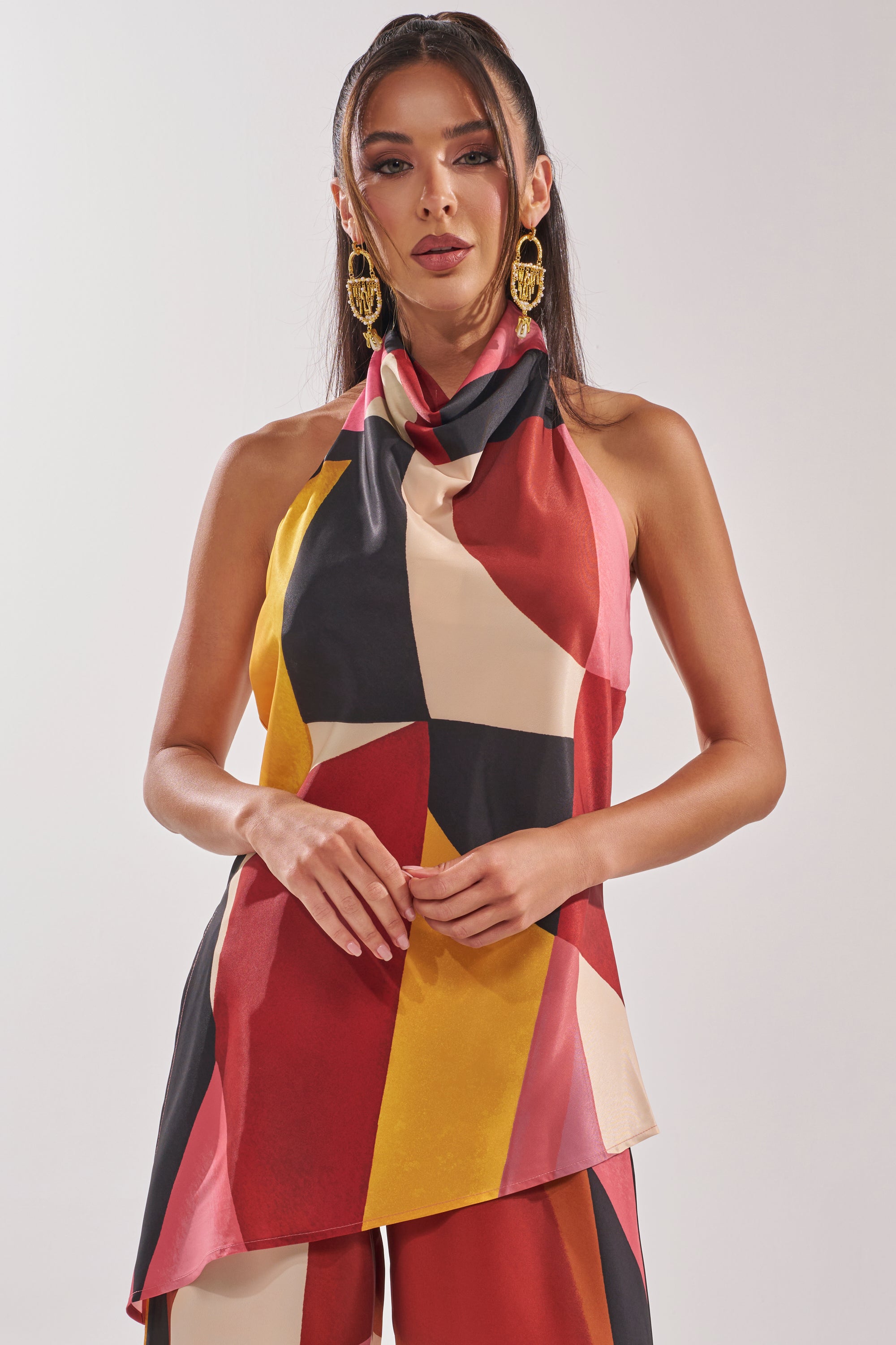 A woman with long dark hair and gold earrings models the MIXED EMOTIONS BLOUSE, a sleeveless top with a multicolored abstract geometric design and draped neckline, standing before a plain light background.