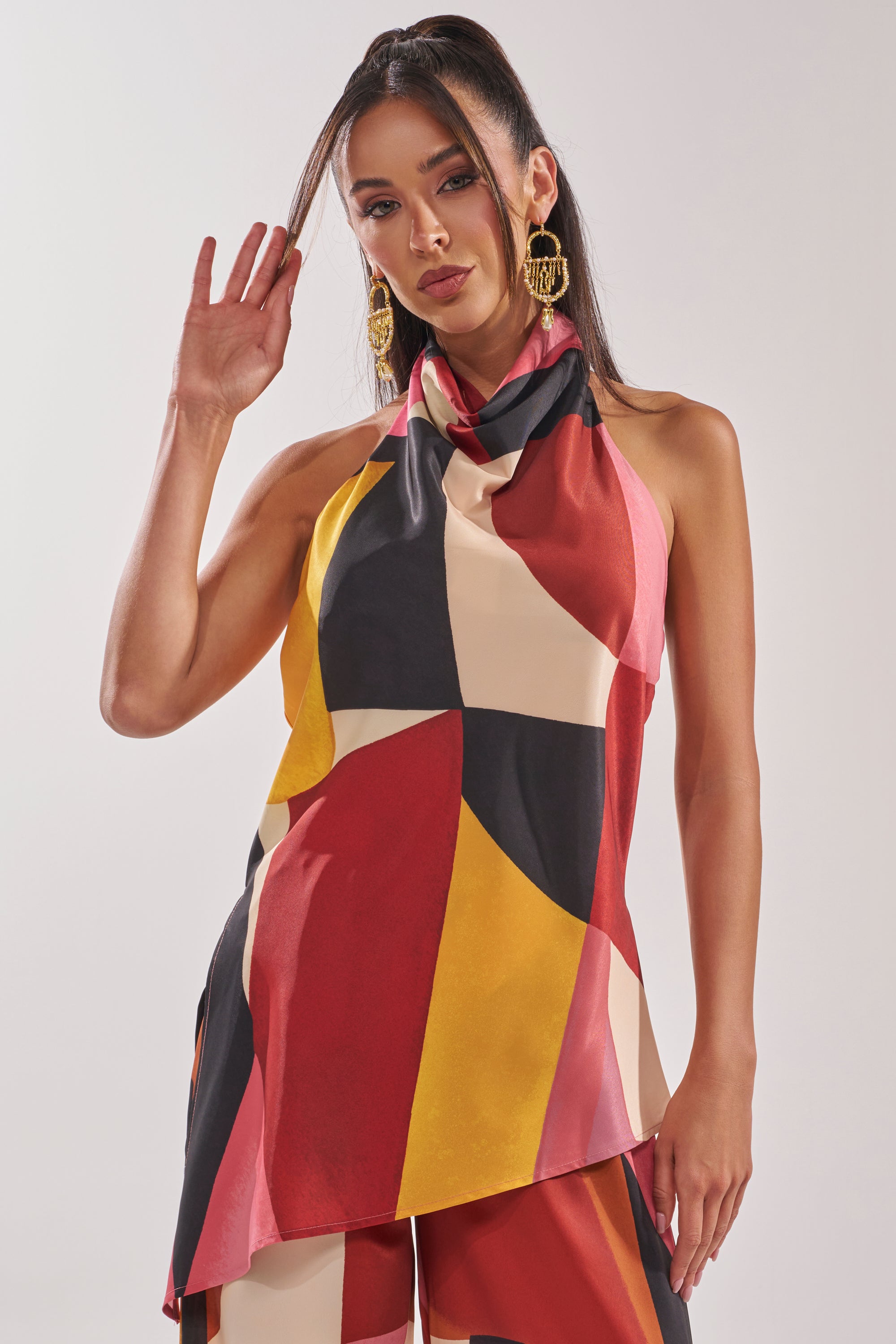 A woman poses against a plain background, wearing the MIXED EMOTIONS BLOUSE—a sleeveless geometric print top—with matching pants and gold earrings, holding one hand up near her face.