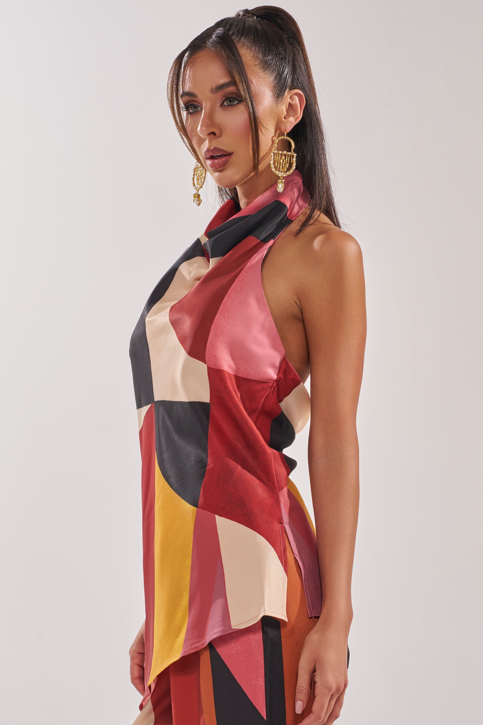 A woman with long, dark hair in a high ponytail wears the MIXED EMOTIONS BLOUSE, featuring a geometric print and sleeveless design, paired with large gold hoop earrings against a plain light background.