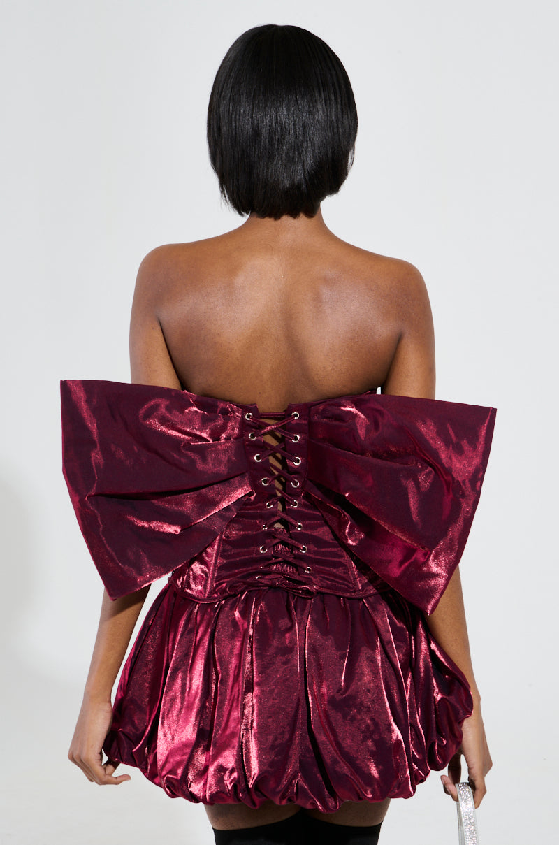 A person with short black hair is turned away, dressed in the Ruby Red Corset Top With Bow Detail. The metallic fabric showcases an oversized bow and corset-style lacing at the back, accompanied by a voluminous skirt. The background is plain and light-colored.