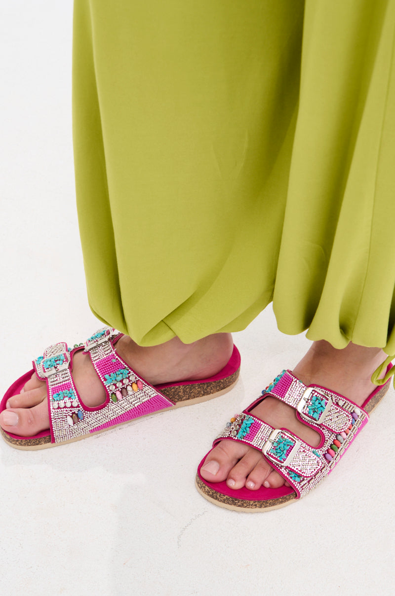 A person wearing bright green loose pants and AKIRA RUVY FUCHSIA STONE EMBELLISHED FLAT SANDAL stands on a white surface.