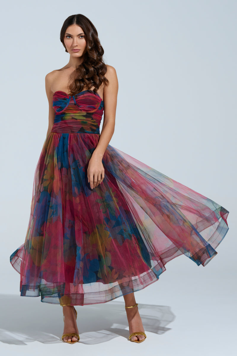Wearing the vibrant floral MEGAN STRAPLESS MAXI DRESS, the woman poses elegantly against a light background. Her long wavy hair cascades gracefully as she dons strappy gold heels, radiating a sophisticated and fashionable allure.