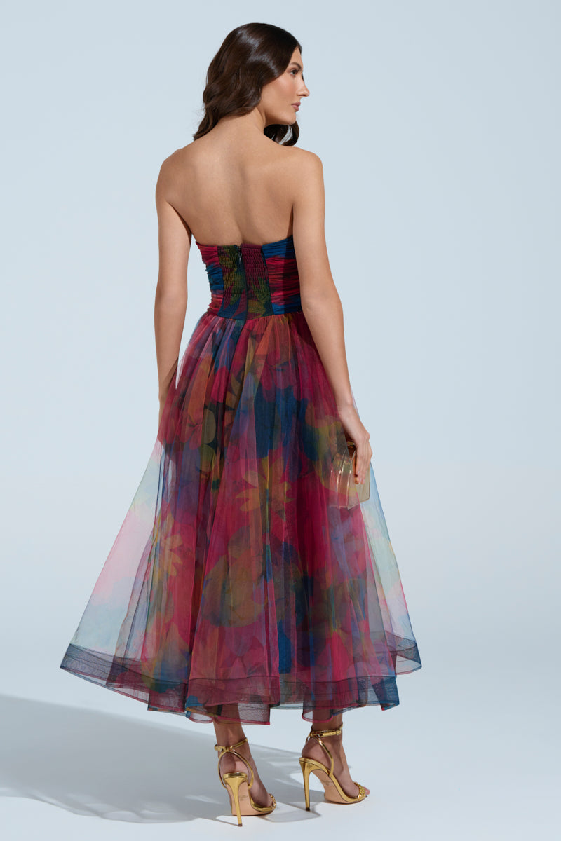 A woman in the MEGAN STRAPLESS MAXI DRESS, featuring a colorful, floral strapless design with a flared, layered skirt. She holds a small clutch and wears gold high-heeled sandals, with her hair styled in loose waves against a simple light blue backdrop.