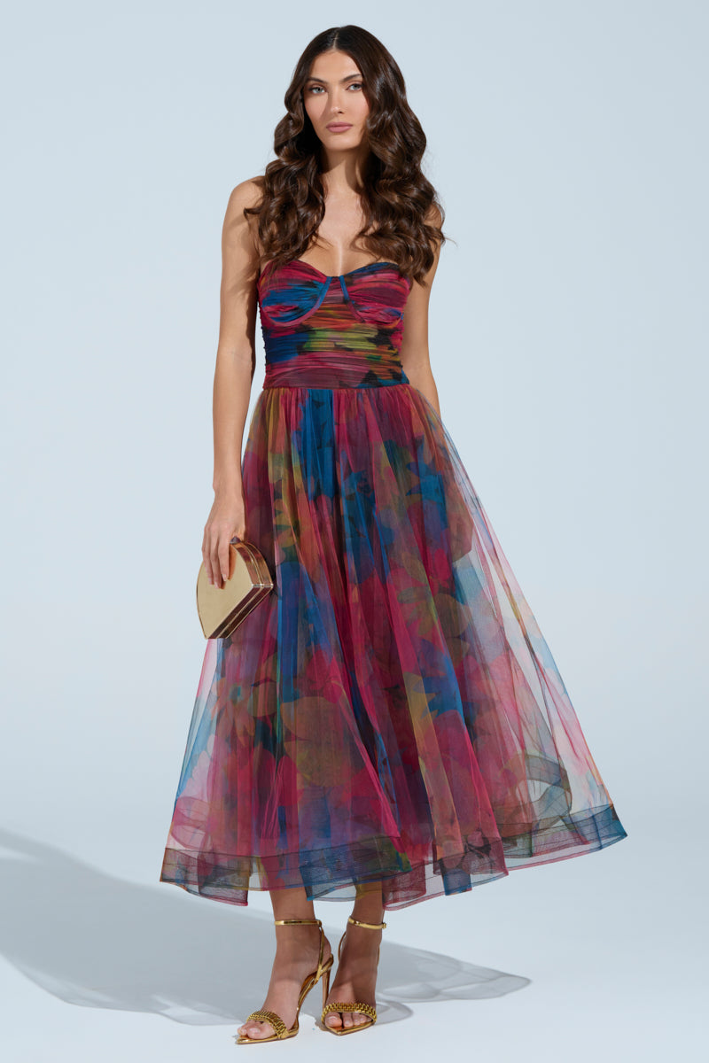 A woman in a colorful MEGAN STRAPLESS MAXI DRESS with a fitted bodice and flared skirt stands against a light blue background. Her long, wavy brown hair flows over her shoulders, perfectly complementing her gold sandals and clutch and enhancing the dress's floral pattern.