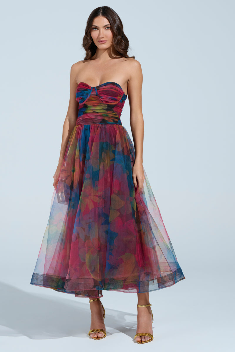 A woman wears the MEGAN STRAPLESS MAXI DRESS, featuring a tulle mesh fabric with a fitted bodice and flowing skirt in vibrant floral hues. She pairs it with strappy high-heeled sandals against a plain background.