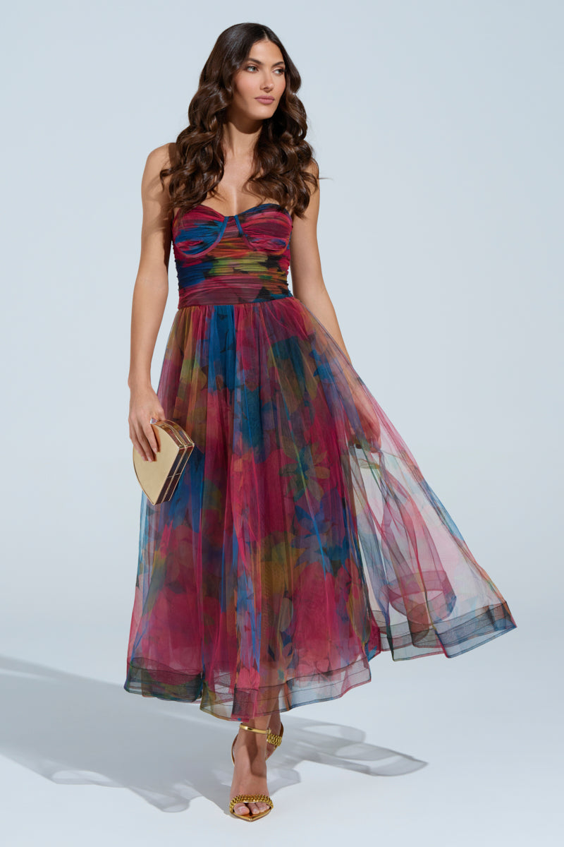 A woman in the MEGAN STRAPLESS MAXI DRESS, featuring a colorful floral pattern, poses against a plain background. She holds a small beige clutch and wears gold heels. Her long, wavy hair cascades down her shoulders as her dress flows elegantly.