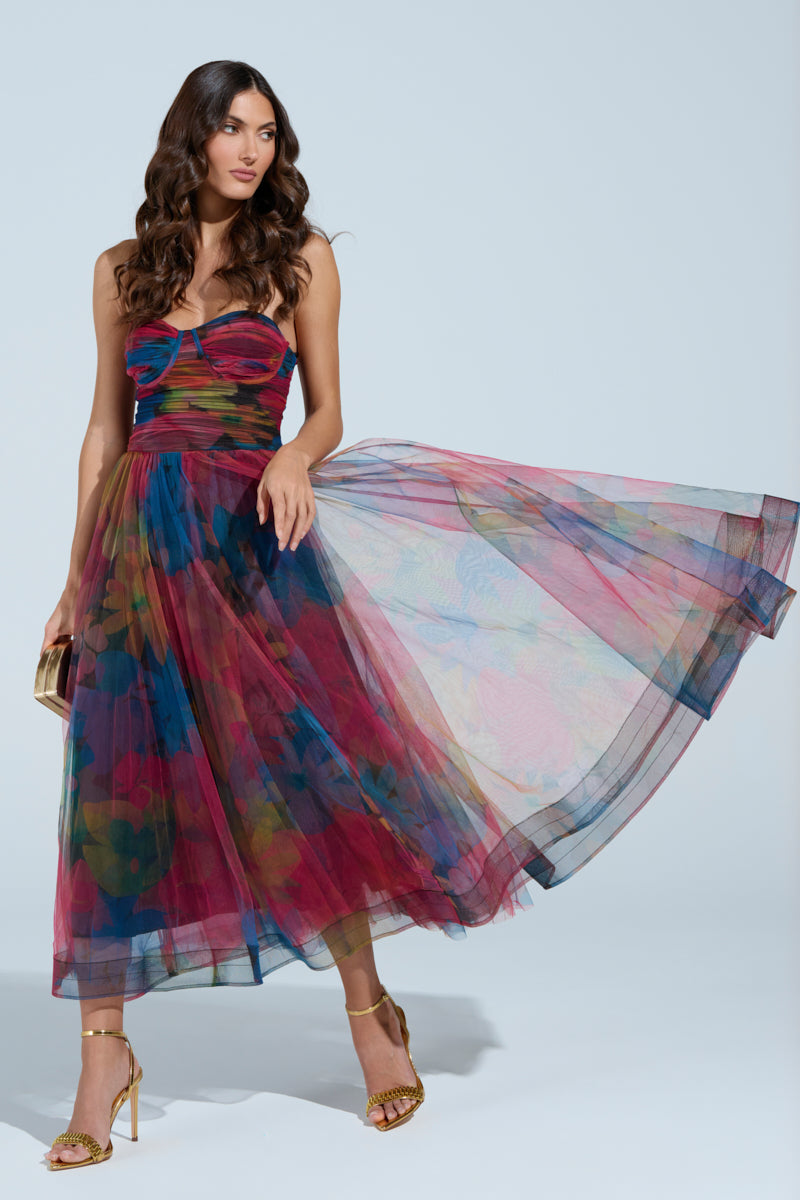 A woman in the MEGAN STRAPLESS MAXI DRESS, featuring a floral pattern and fitted bodice, poses against a plain background. Her long, wavy hair flows over her shoulders as she holds a clutch and wears gold heels.