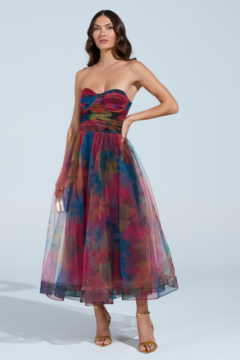 A woman poses confidently in the MEGAN STRAPLESS MAXI DRESS featuring a vibrant floral pattern and tulle mesh. Her loose, wavy hair enhances the elegance of her gold high-heeled sandals against a plain, light-colored background.