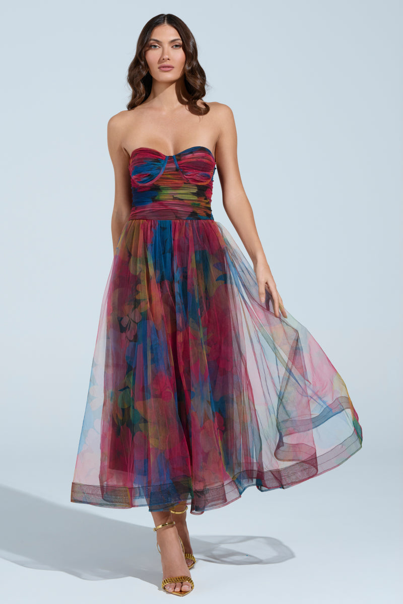 A woman in the MEGAN STRAPLESS MAXI DRESS, featuring a multicolored floral pattern with a fitted bodice and flowing skirt, stands against a light blue background. With long brown hair and strappy gold heels, she exudes elegance and charm.