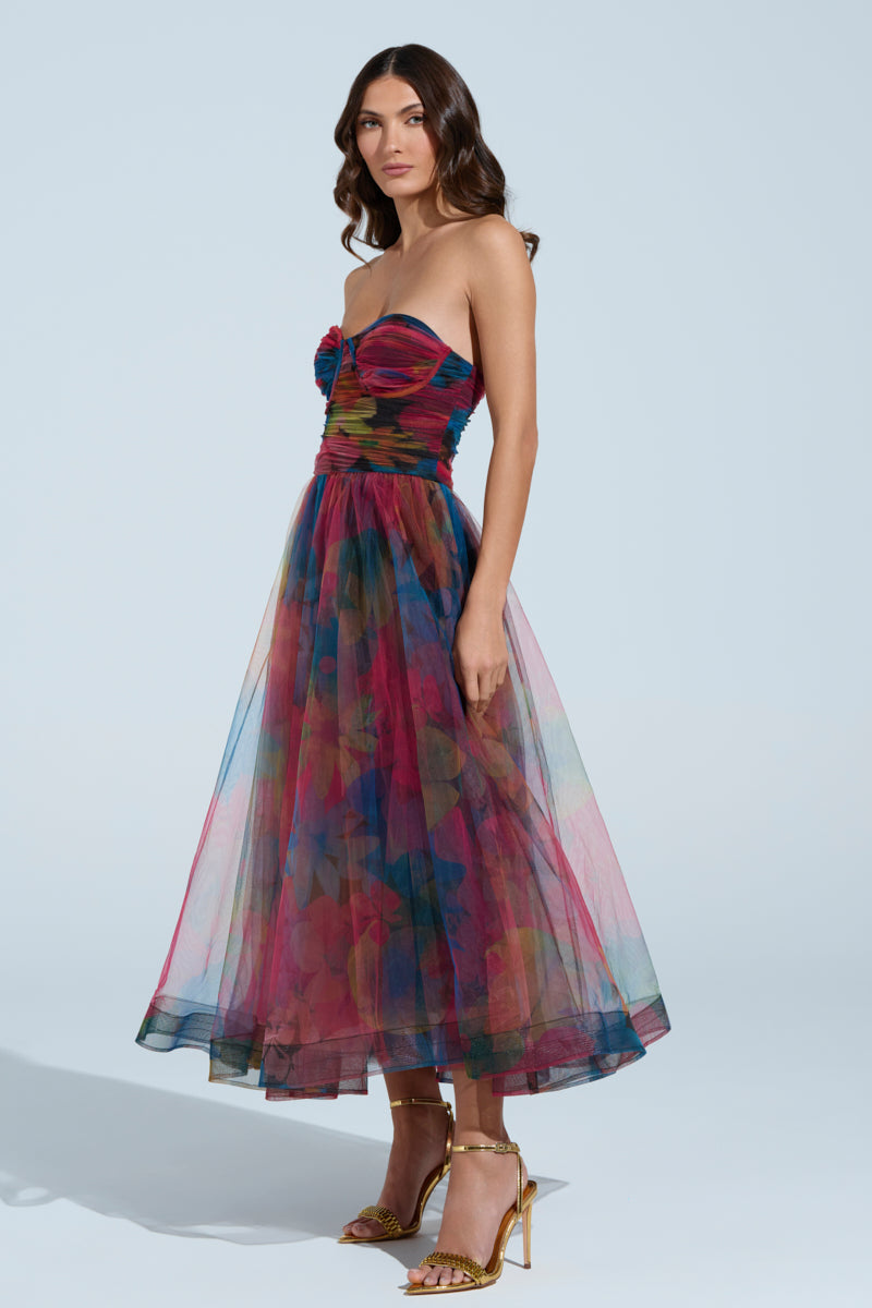 In the MEGAN STRAPLESS MAXI DRESS, a woman showcases a vibrant floral pattern on light blue. The tulle mesh creates a fitted bodice and full skirt. Gold strappy high heels enhance her style as her dark hair falls in loose waves.