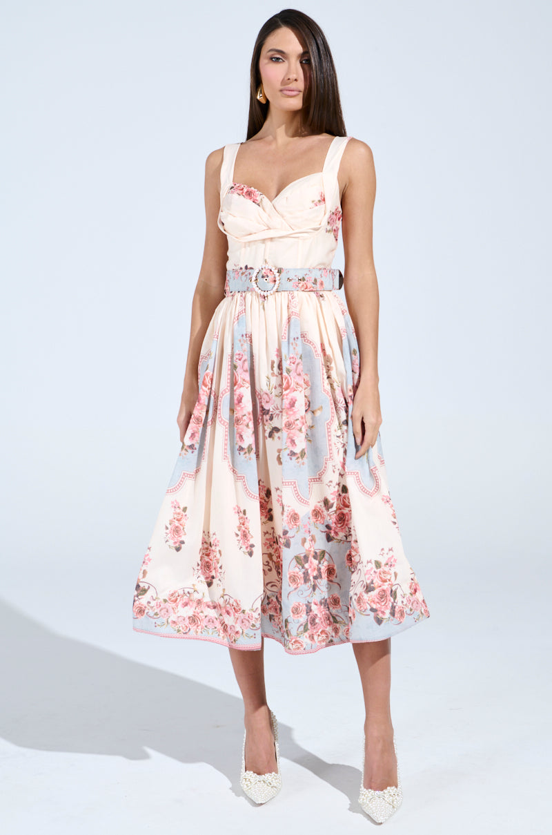 A woman poses in the HANNA FLORAL MIDI DRESS, featuring a vintage-inspired design with pleated skirt and fitted bodice adorned in pink and blue patterns. She pairs it with white high heels, her long brown hair standing out against the plain white backdrop.