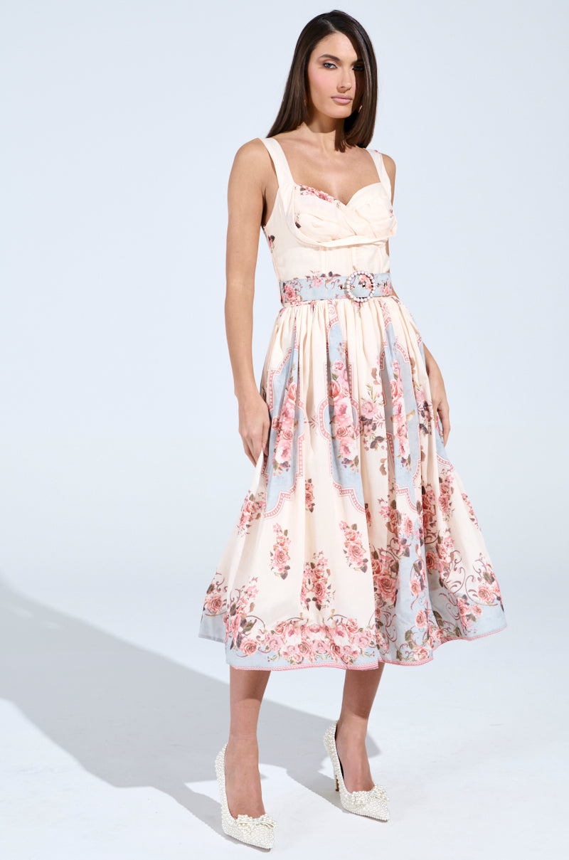 Wearing the HANNA FLORAL MIDI DRESS with a corset-like bodice and a flared skirt featuring pink and blue flowers on a light background, a woman elegantly pairs it with white high-heeled shoes against a plain backdrop.