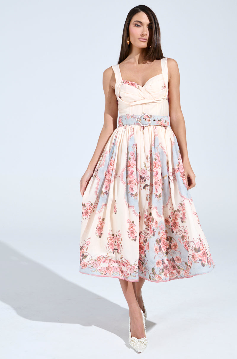 A woman models the HANNA FLORAL MIDI DRESS, showcasing its vintage-inspired design with a corset-like bodice and flared skirt in pink and blue hues. She accessorizes with white heels, her long straight hair contrasting against a simple backdrop.