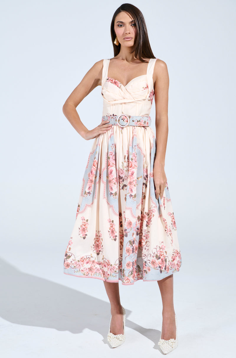 A woman exudes confidence in the HANNA FLORAL MIDI DRESS, featuring a vintage-inspired design and belted waist. Her long hair pairs beautifully with her neutral makeup and white textured high heels, all set against a plain white background that highlights the elegance of her ensemble.