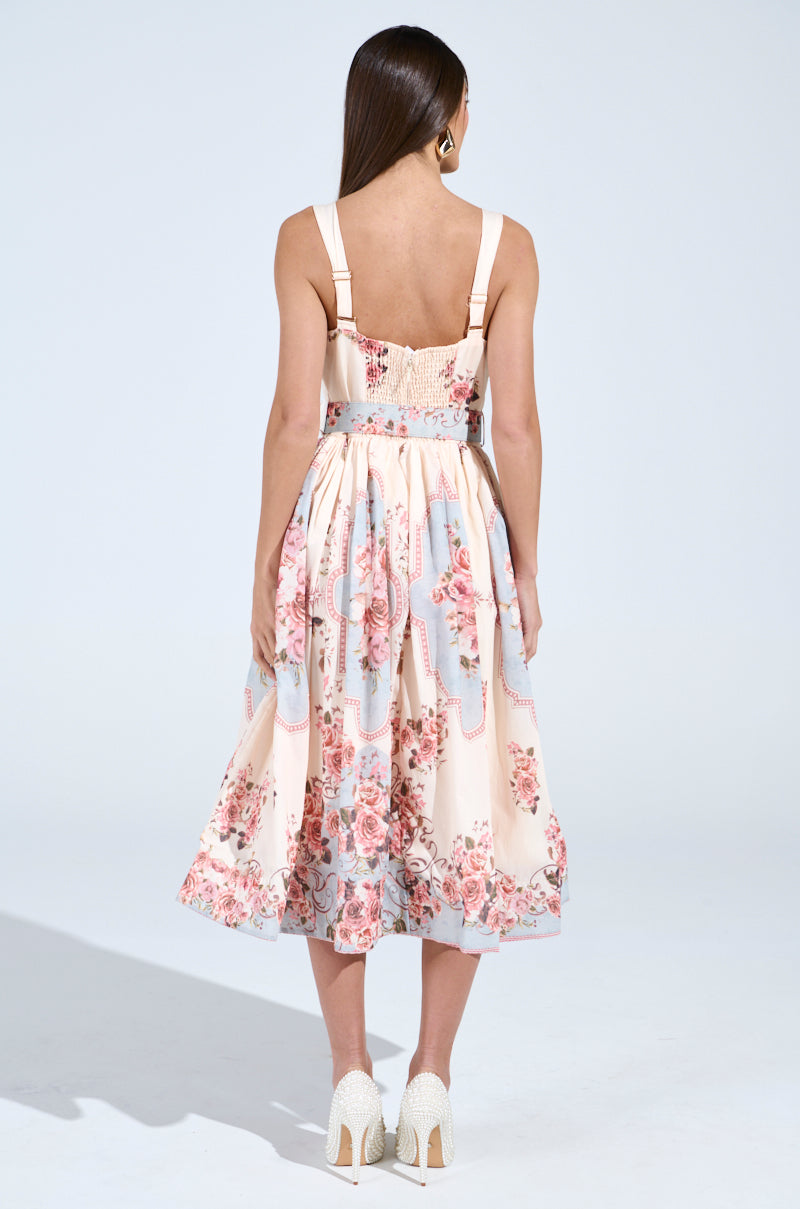 A woman with long brown hair faces away, wearing the sleeveless HANNA FLORAL MIDI DRESS that showcases vintage-inspired floral patterns and lace detailing. The light dress features pink and blue flowers, paired with high-heeled white shoes.