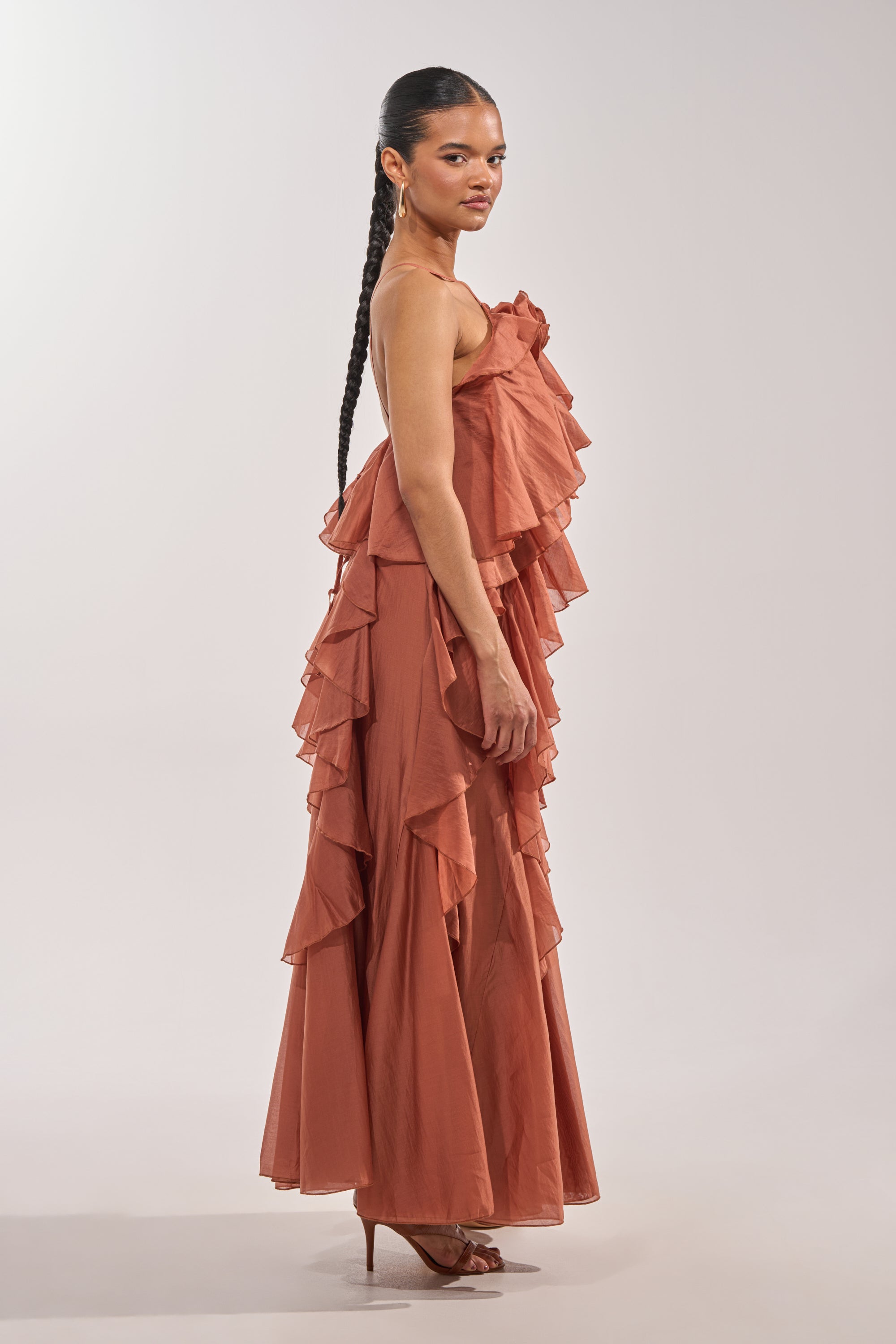 A woman in profile models the KISS ME SLOW CHIFFON MAXI DRESS IN RUST, featuring tiered ruffles. She wears heels, her long hair styled in a single braid, and poses against a simple light background. 3