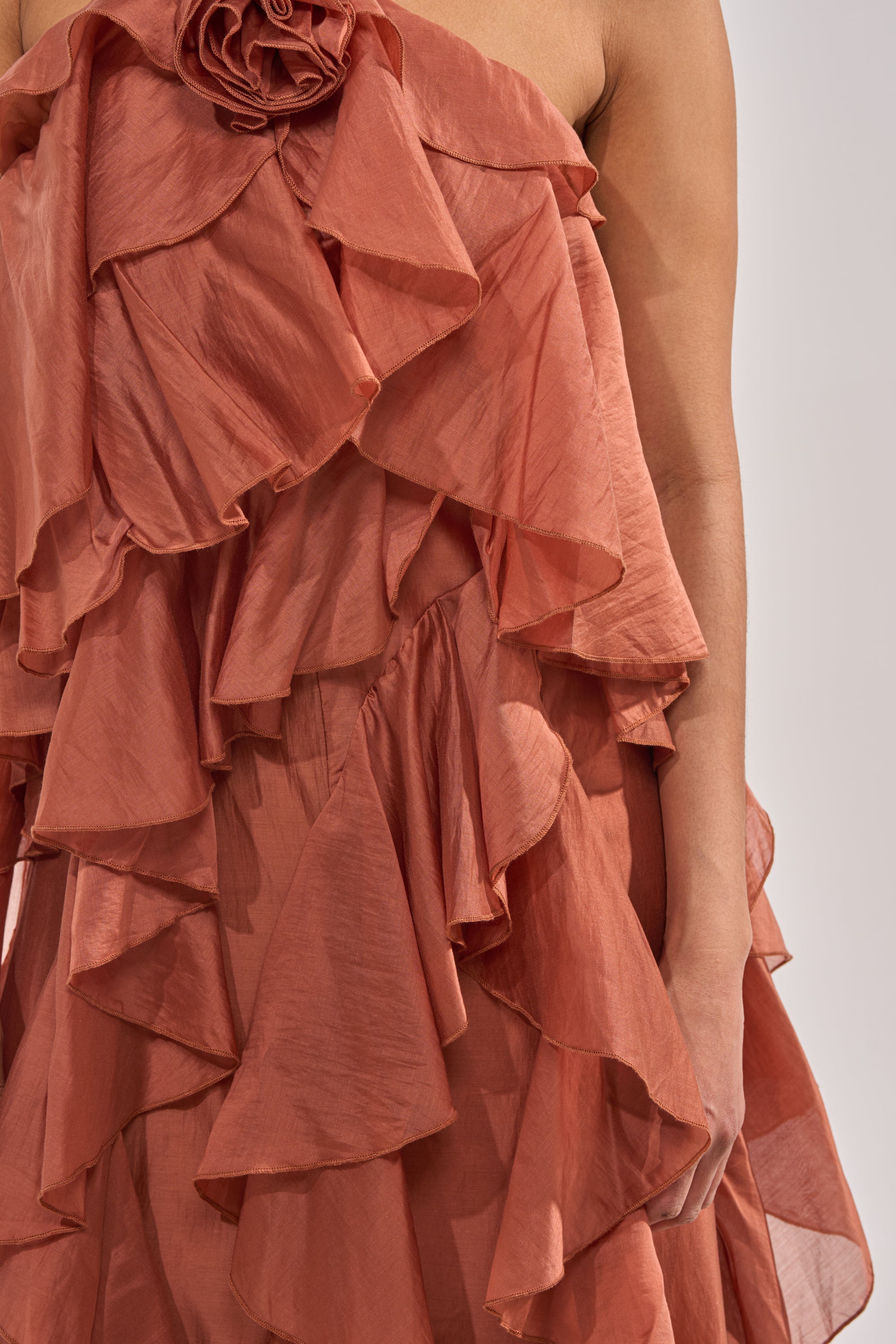 A close-up of a person wearing the KISS ME SLOW CHIFFON MAXI DRESS IN RUST, featuring a strapless design, layered ruffles, and a floral appliqué near the neckline. Only their upper body and one arm are visible. 5