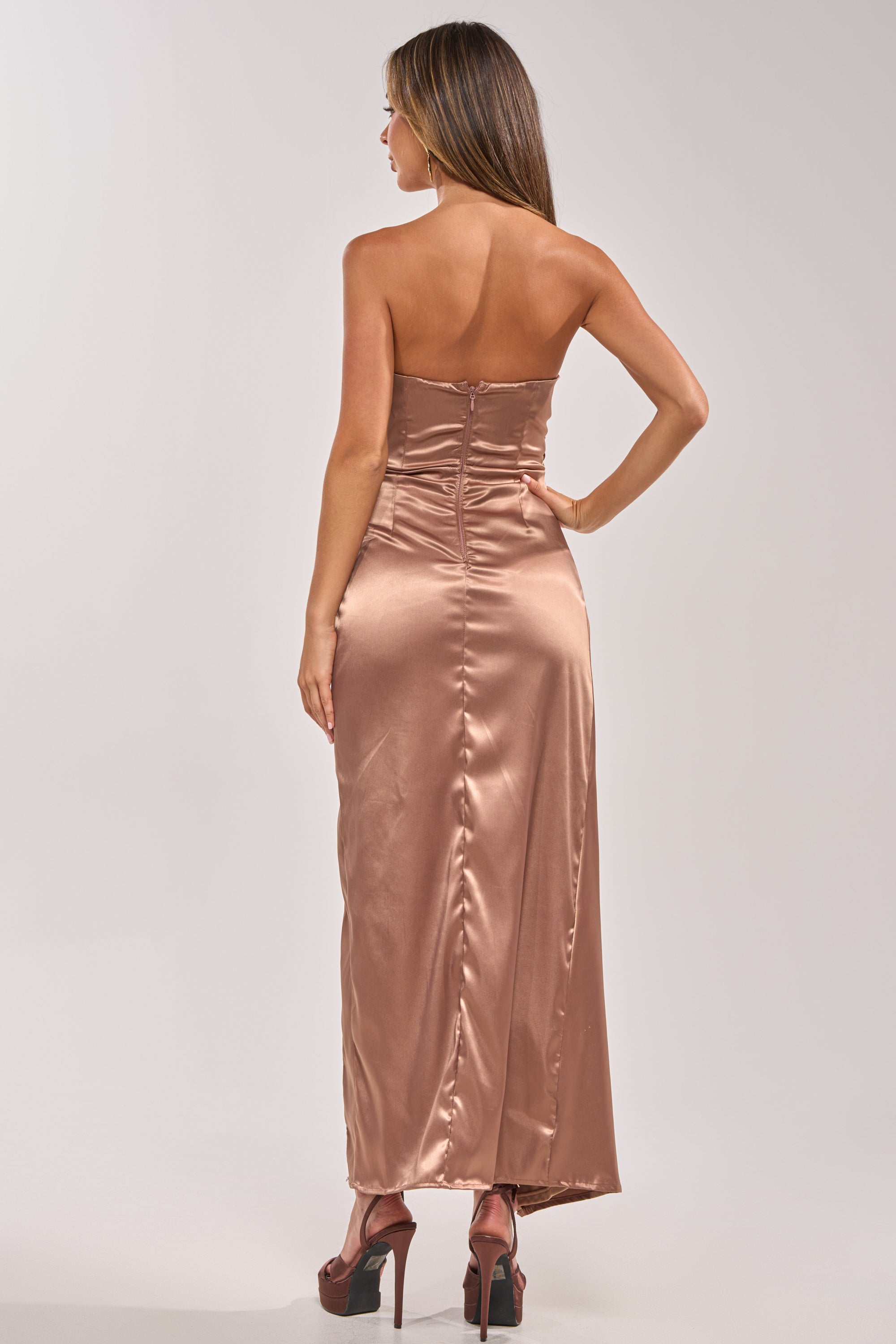 A woman faces away, wearing the HAVING A BALL SATIN MAXI DRESS IN BROWN— a fitted, strapless maxi dress with a ruched bodice, shiny satin finish, visible back zipper, and high heels against a plain light background. 4