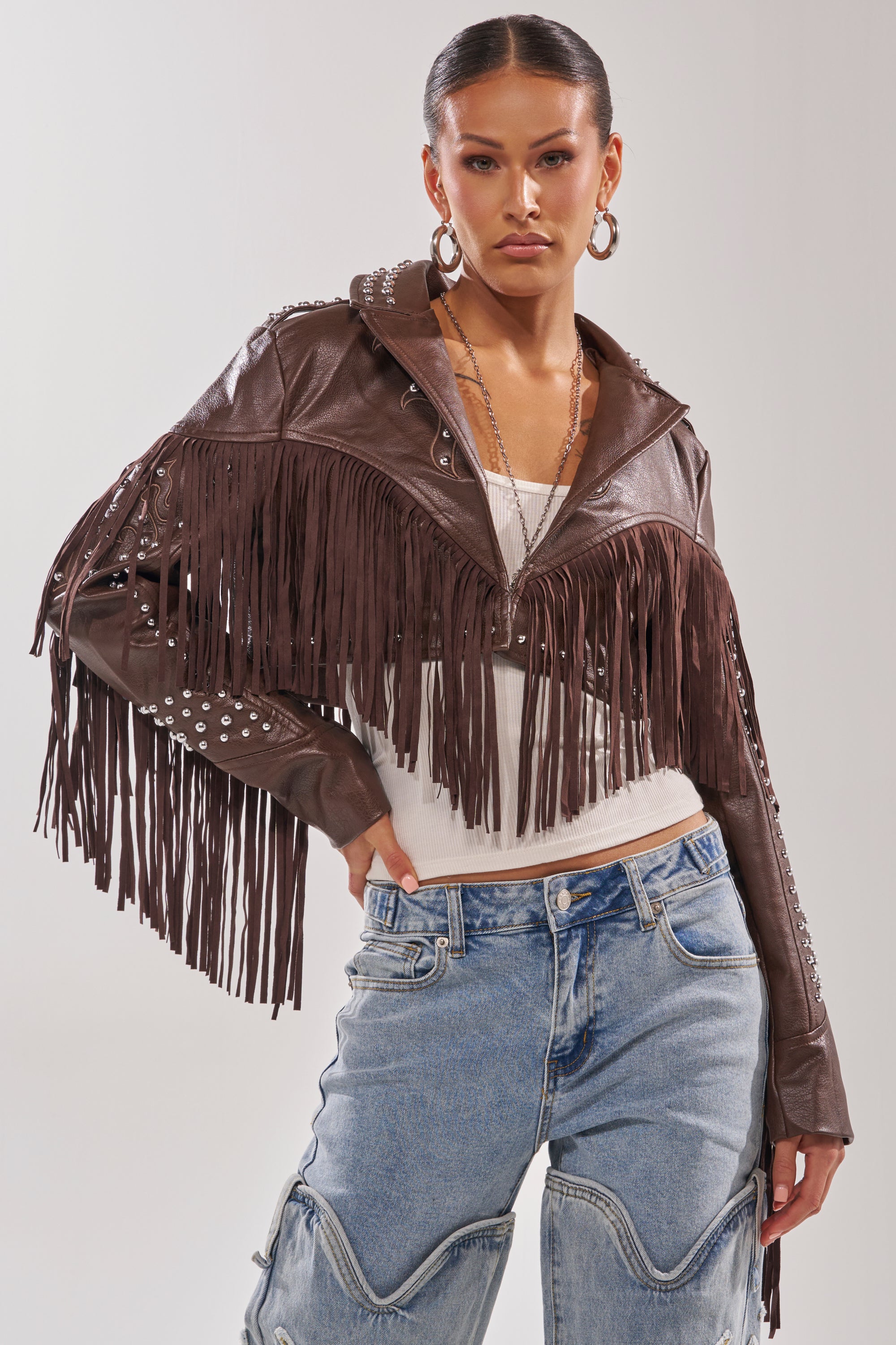 A woman wearing the NOT MY FIRST RODEO CROPPED MOTO—a vintage-inspired western faux leather jacket with fringe and silver studs—pairs it with a white top, large hoop earrings, and light blue jeans, standing confidently against a plain backdrop. 0