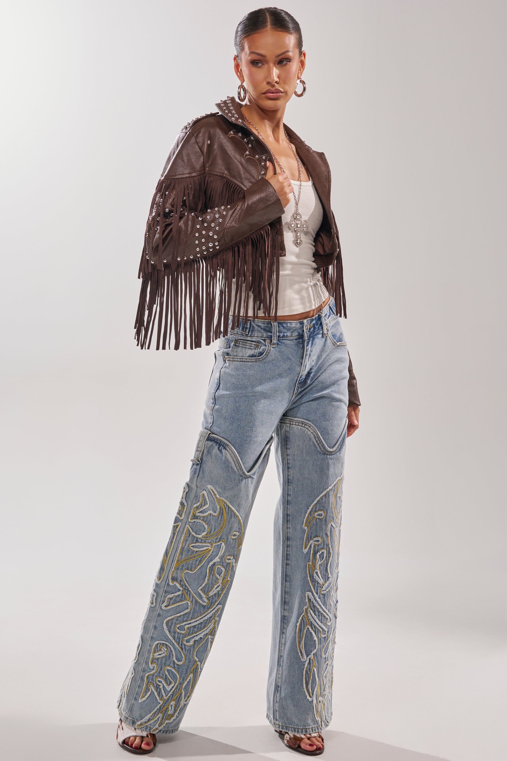 A woman stands confidently in the NOT MY FIRST RODEO CROPPED MOTO jacket, styled with a white top, light blue embroidered wide-leg jeans, hoop earrings, layered necklaces, and open-toe heels. 9