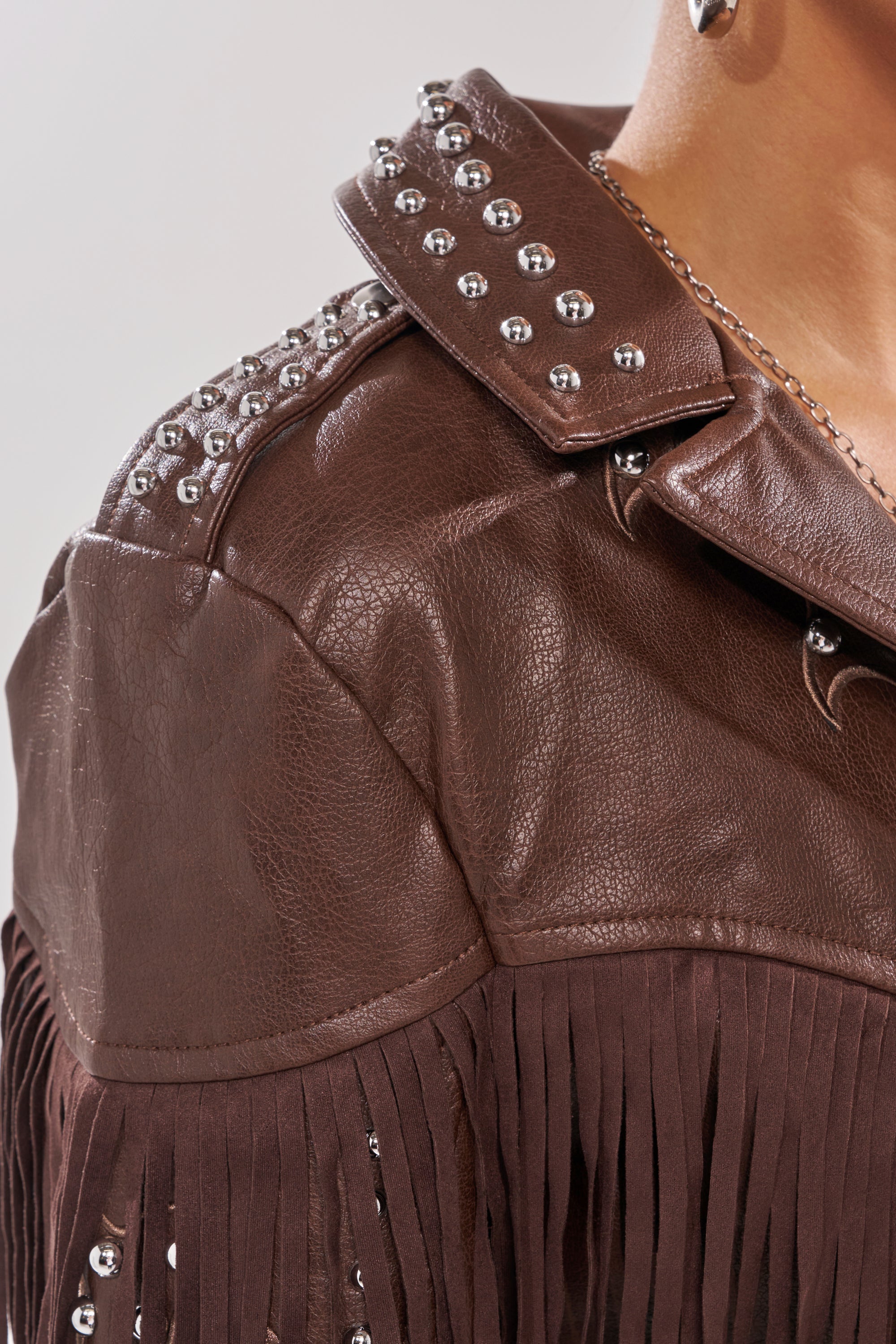 Close-up of a person wearing the NOT MY FIRST RODEO CROPPED MOTO, a western-inspired faux leather jacket featuring silver studs on the collar and shoulder, sleeve fringe detailing, and a visible silver chain necklace. 10