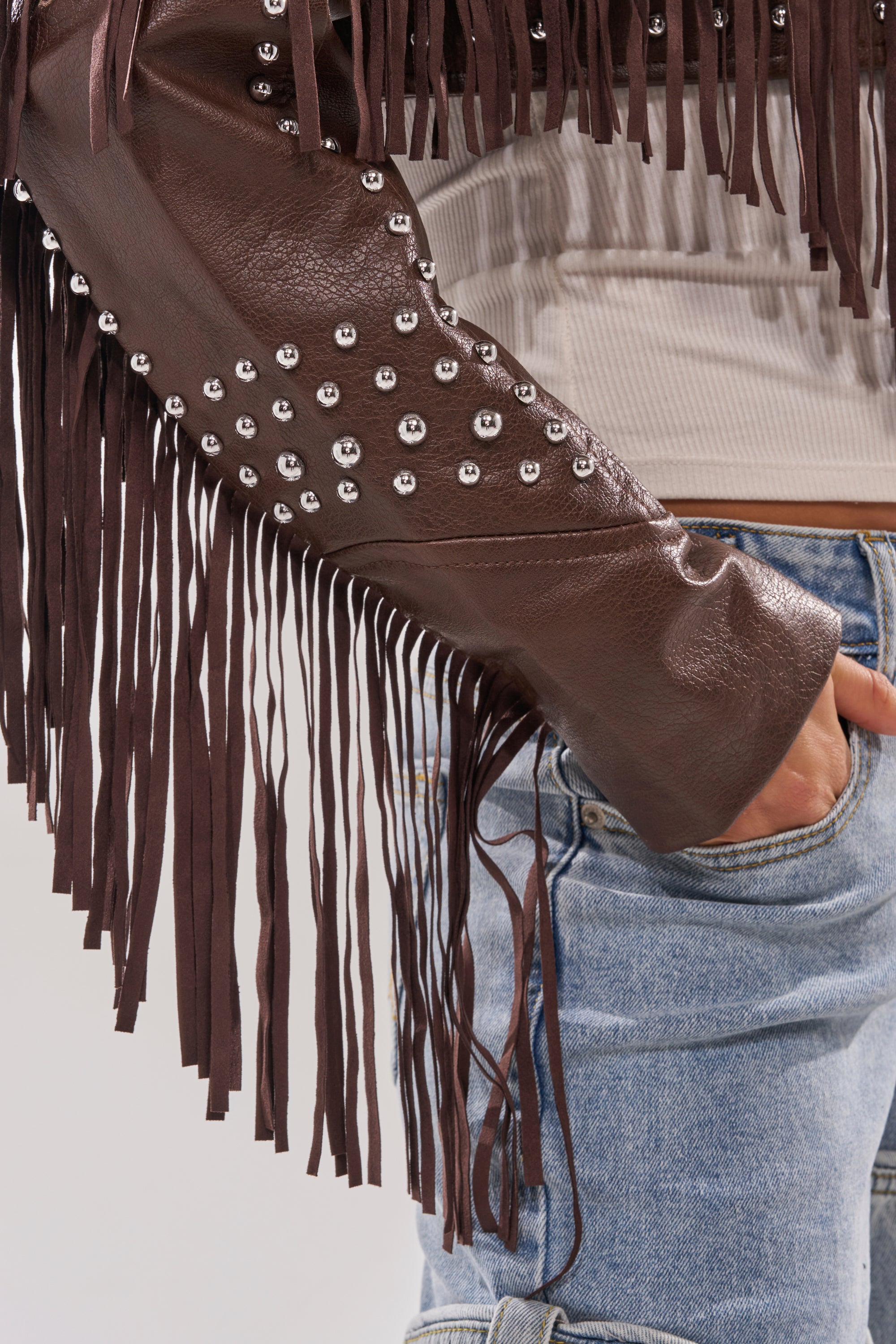 A person wears the NOT MY FIRST RODEO CROPPED MOTO jacket with fringe and silver studs, posing with one hand in the pocket of light blue jeans. The detailed sleeve and fringe of the jacket are in focus. 11