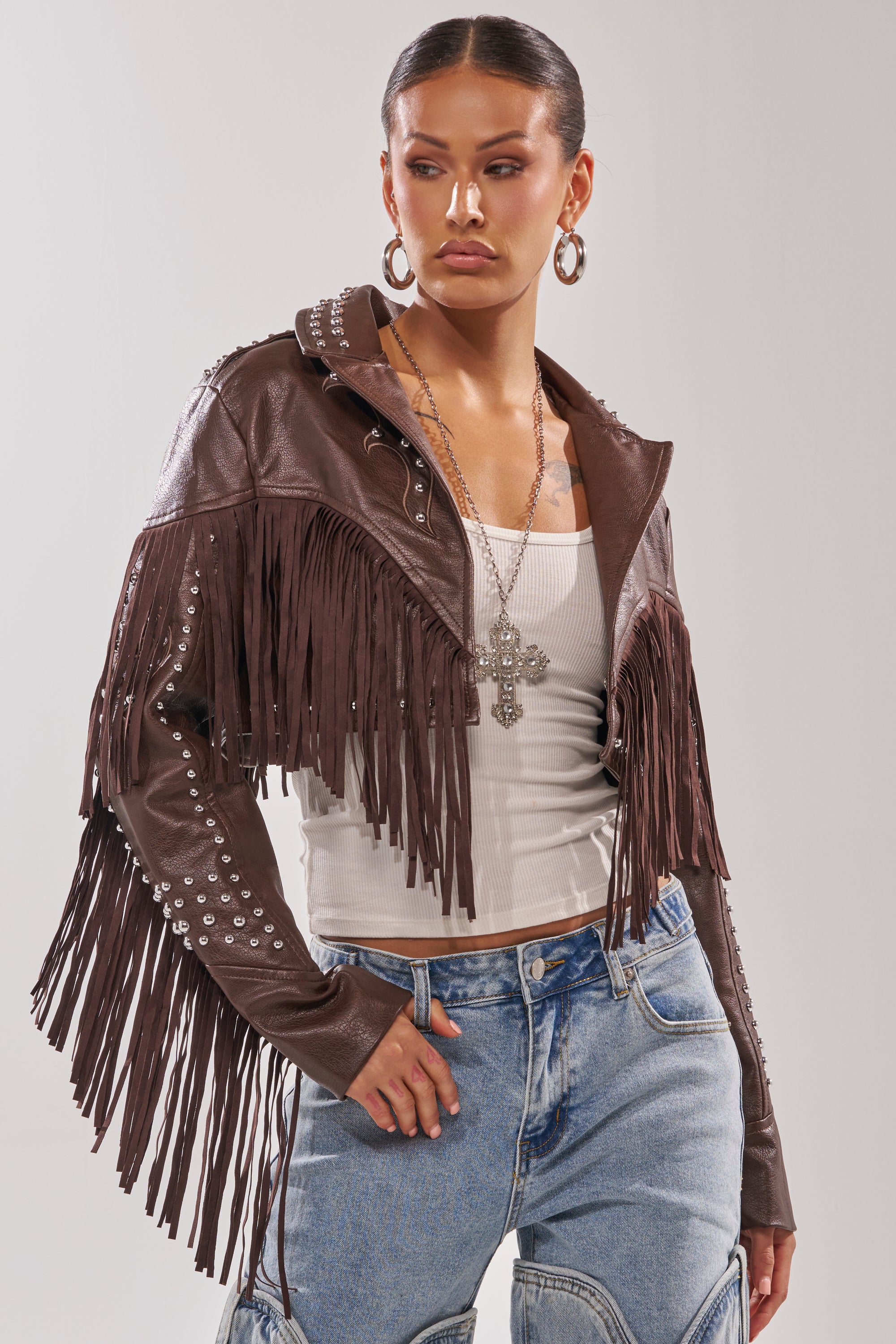 A woman models the NOT MY FIRST RODEO CROPPED MOTO—a brown western fringe jacket with silver studs—paired with a white ribbed tank, layered necklaces, hoop earrings, and blue jeans against a plain background. 1