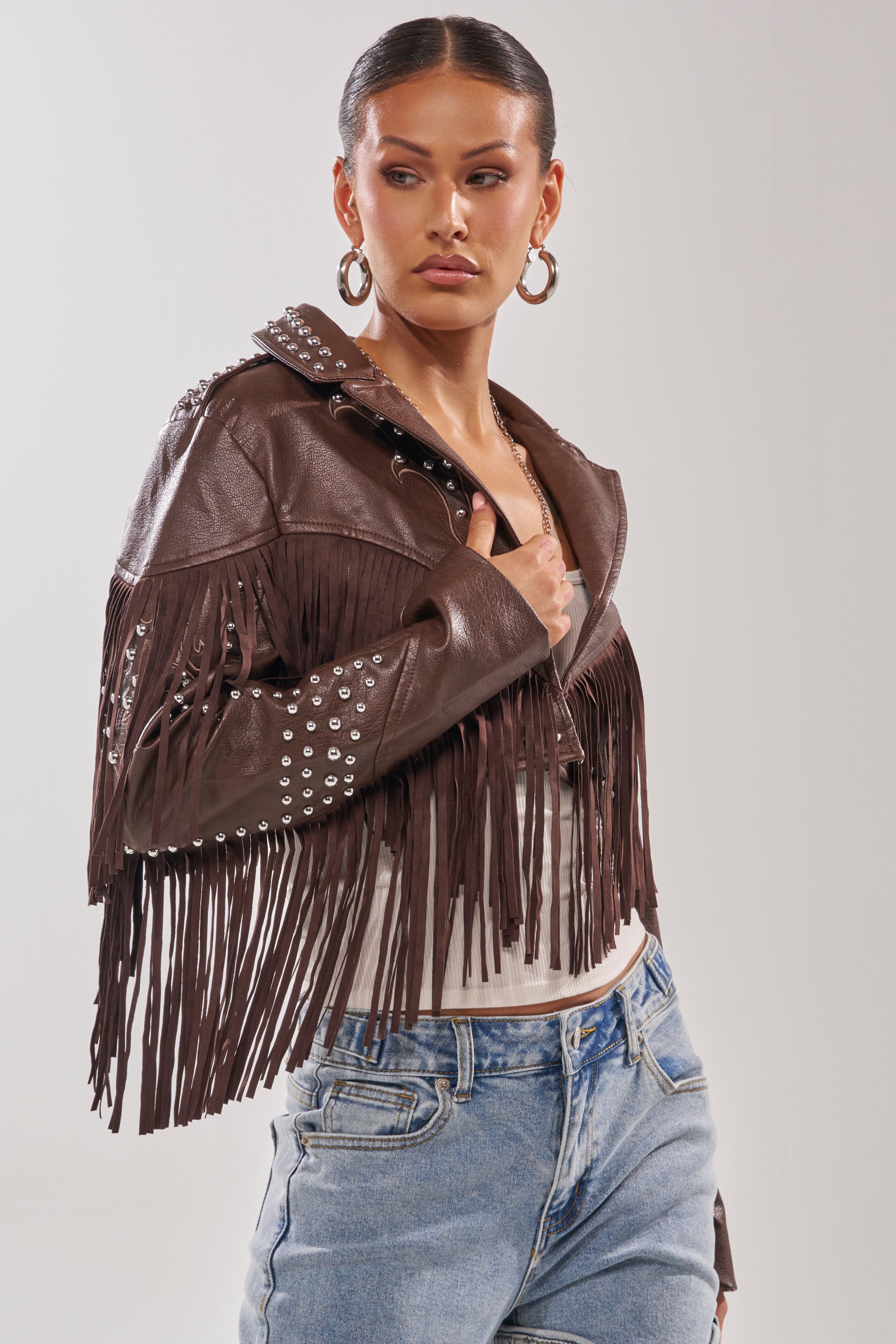 A woman wears the NOT MY FIRST RODEO CROPPED MOTO jacket with silver studs over a white top and blue jeans, looking to the side with a confident expression. 2