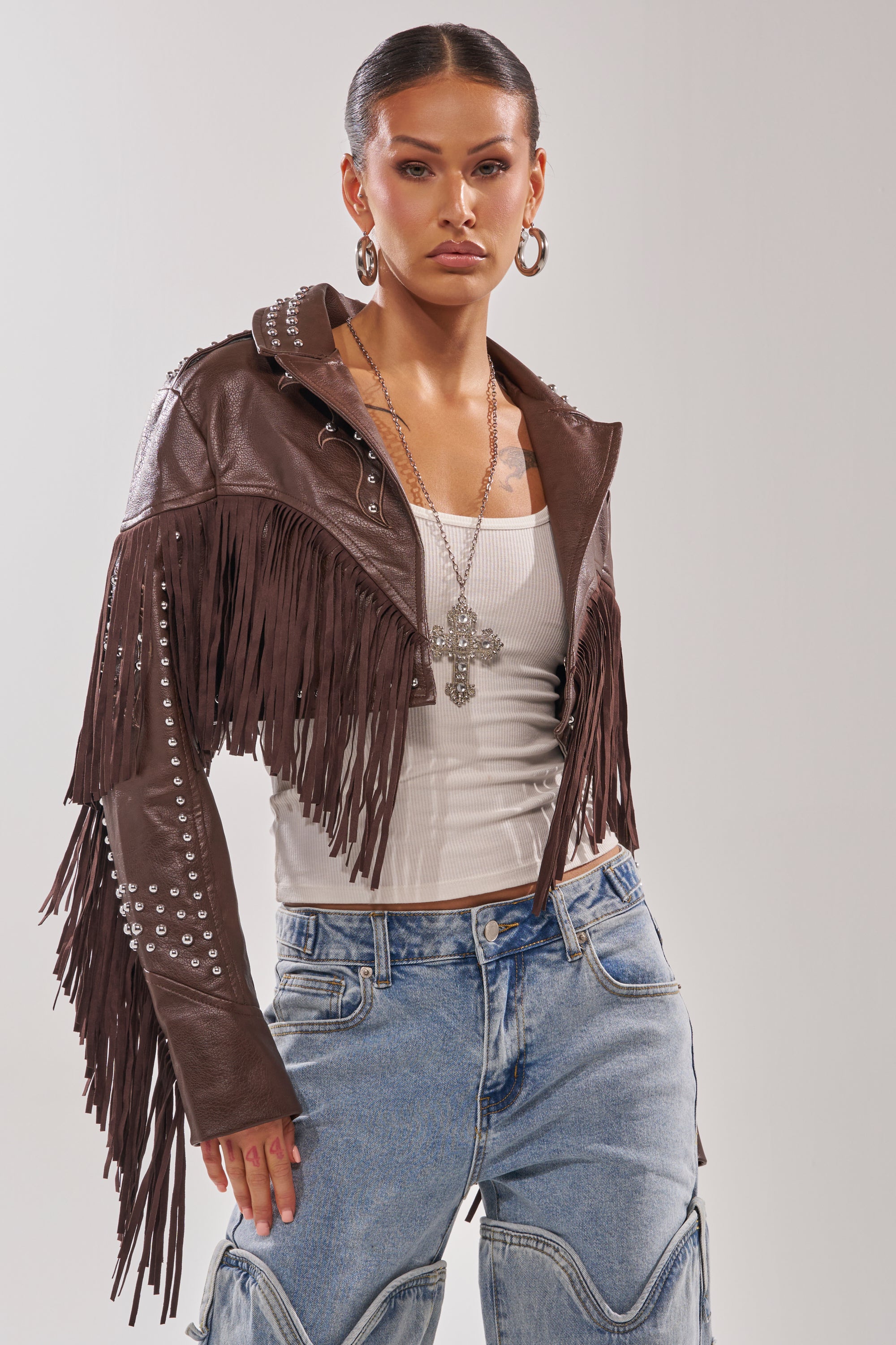 A woman wears the NOT MY FIRST RODEO CROPPED MOTO brown faux leather fringe jacket with silver studs, styled with a white ribbed tank top, layered necklaces, hoop earrings, and loose blue jeans against a plain light background. 3