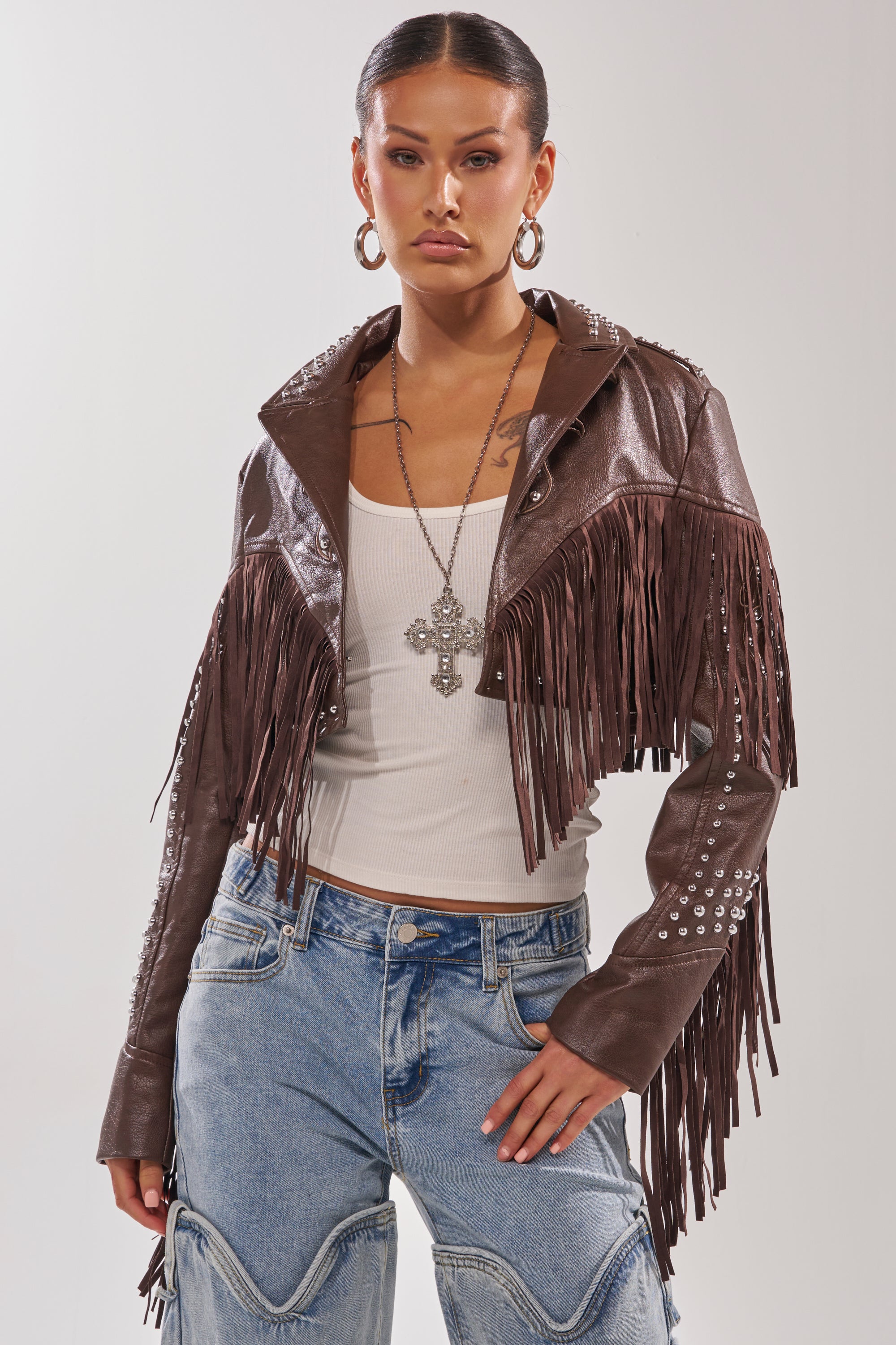 Woman wearing the NOT MY FIRST RODEO CROPPED MOTO brown western fringe leather jacket with metal studs over a white ribbed tank and blue jeans, accessorized with hoop earrings and a long cross necklace. 4