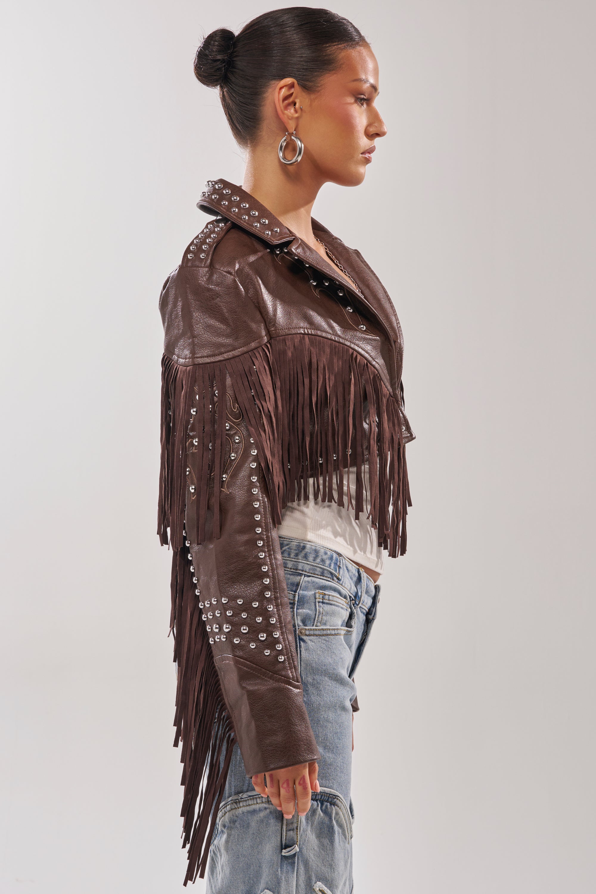 A woman in profile wears the NOT MY FIRST RODEO CROPPED MOTO jacket with fringe and studs, paired with light-wash ripped jeans and large hoop earrings, her hair in a bun, standing against a plain light background. 5