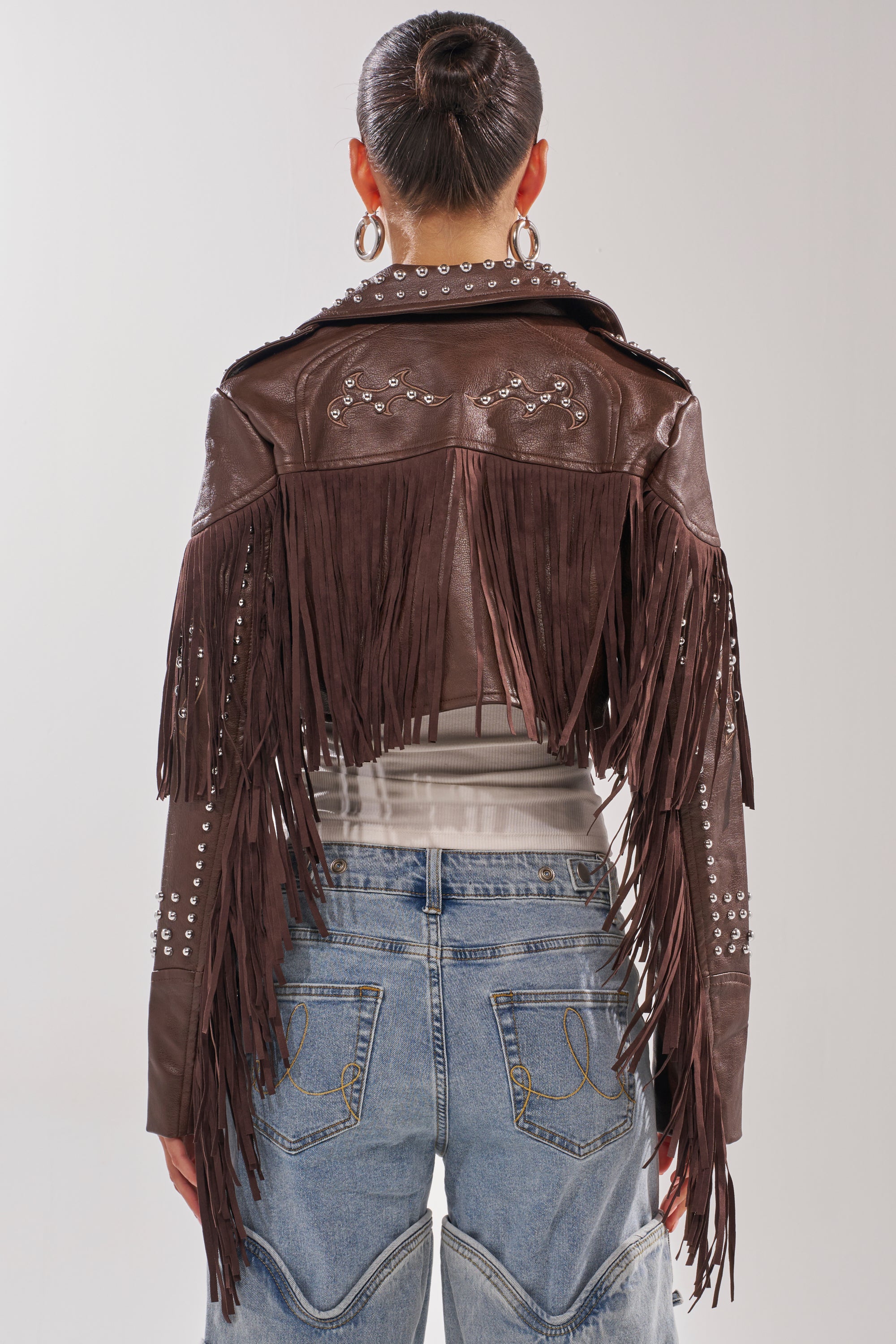 A person stands facing away, wearing the NOT MY FIRST RODEO CROPPED MOTO—a brown western jacket with fringe and stud details—paired with large hoop earrings, blue jeans with unique stitching, and hair styled in a neat bun. 6