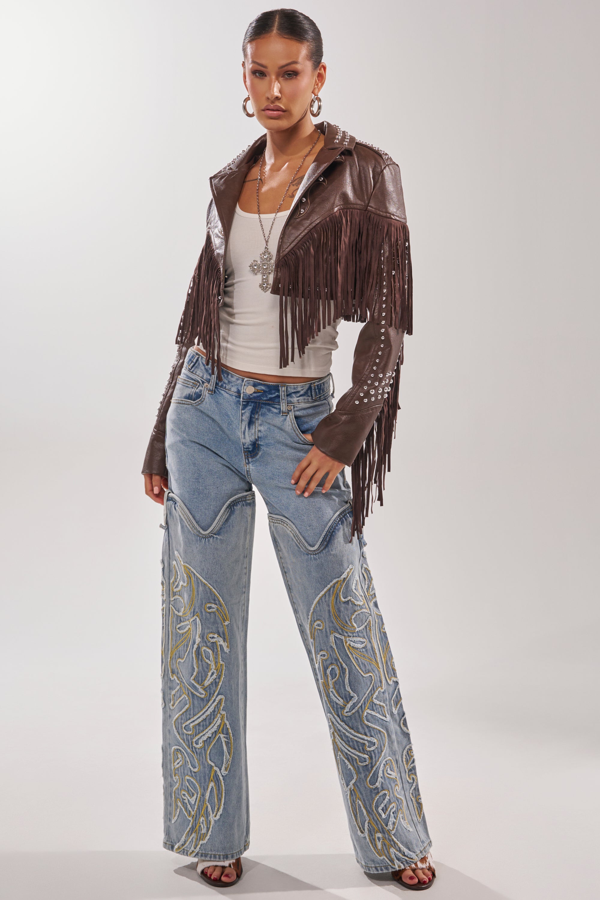 A woman poses confidently in the NOT MY FIRST RODEO CROPPED MOTO faux leather fringe jacket over a white tank top, embroidered wide-leg jeans, hoop earrings, a necklace, and heeled sandals against a plain background. 7