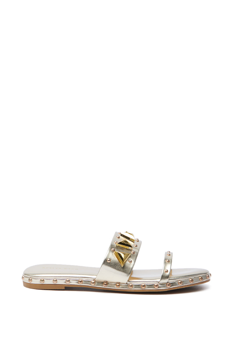 The AKIRA RYLIE GOLD EMBELLISHED SLIP ON SANDAL features a metallic faux leather finish with two slender straps, small studs, a central decorative element, and a flat sole. It's elegantly displayed against a plain white background.