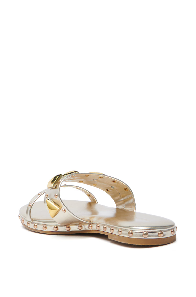 The AKIRA RYLIE GOLD EMBELLISHED SLIP ON SANDAL features a stylish design with metallic faux leather straps, metallic studs, and a flat sole. A light-colored base with gold and silver detailing completes its elegant appearance.