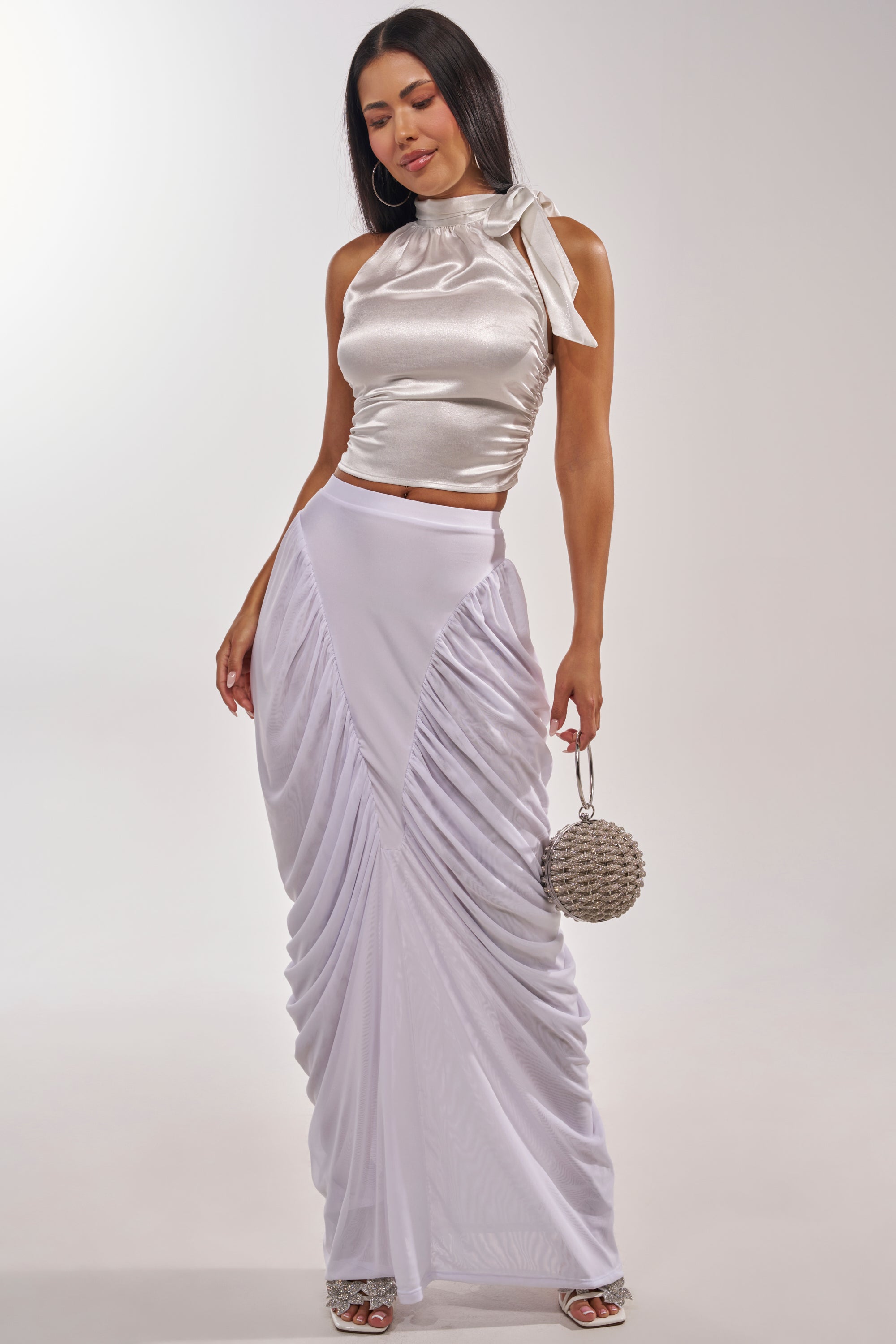 A woman wears the DREAMY RUCHED SKIRT IN WHITE with a shiny silver sleeveless top and tied neck, holding a round beaded purse while smiling on a plain, light background. 0