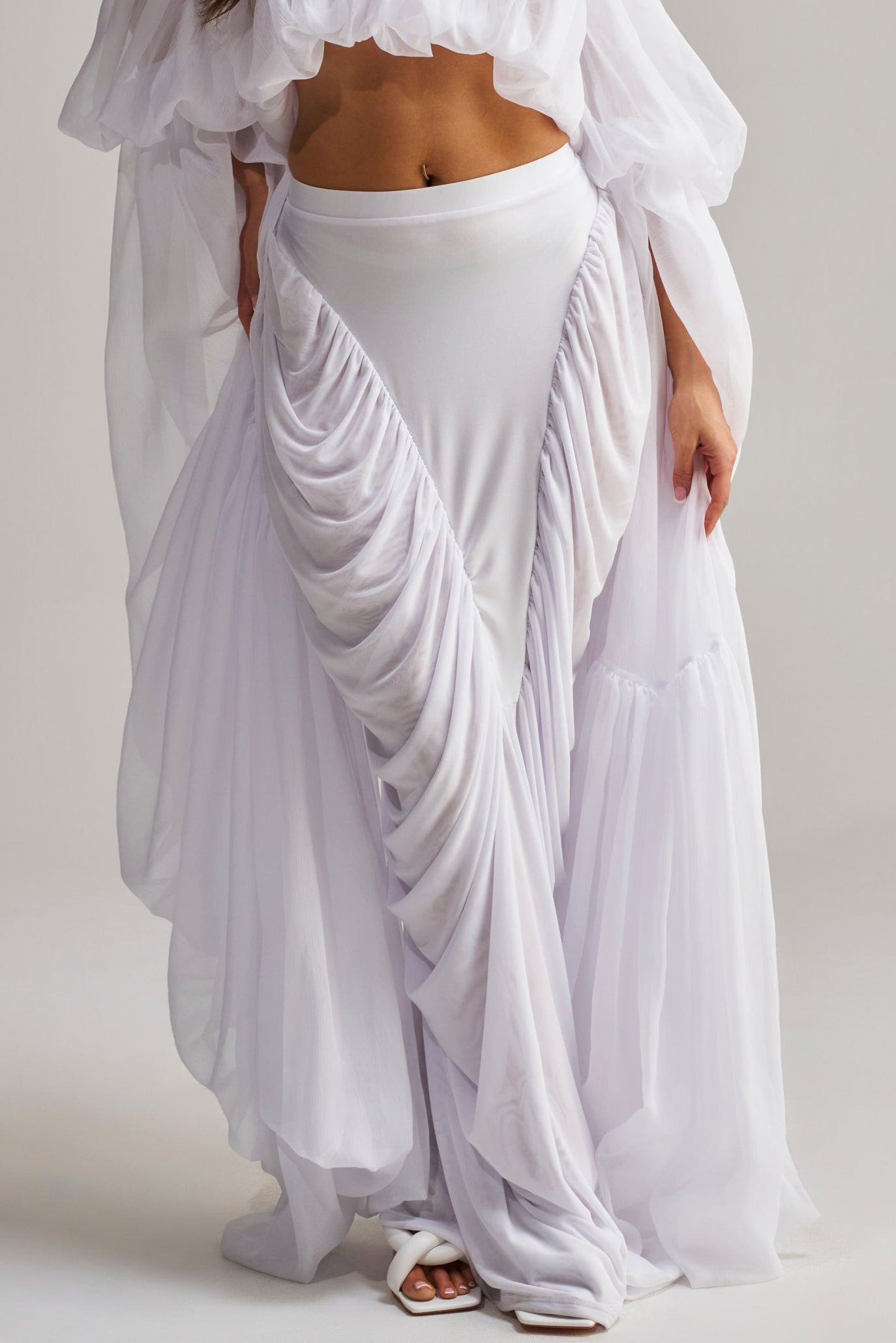 A woman stands facing forward against a plain light background, wearing a flowing white off-shoulder crop top and the DREAMY RUCHED SKIRT IN WHITE, accessorized with a floral necklace.