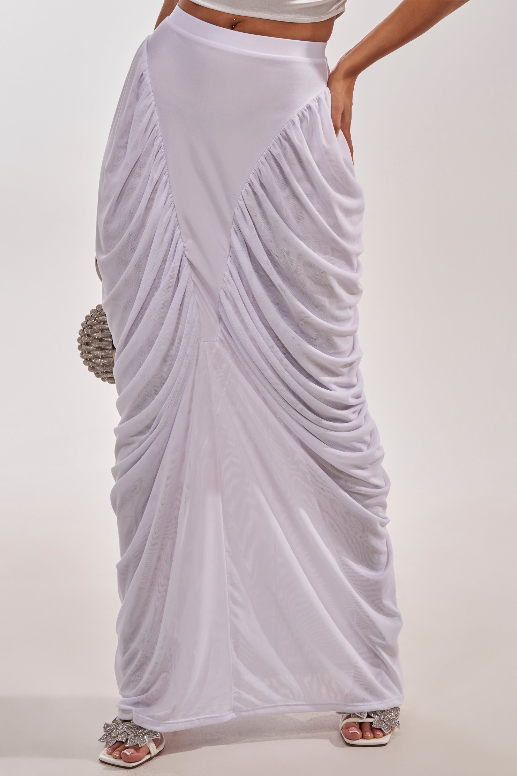 A person wears the DREAMY RUCHED SKIRT IN WHITE, featuring a high-rise fit, ruched sides, and a triangular center panel, paired with white sandals and a beaded clutch against a plain light background. 1