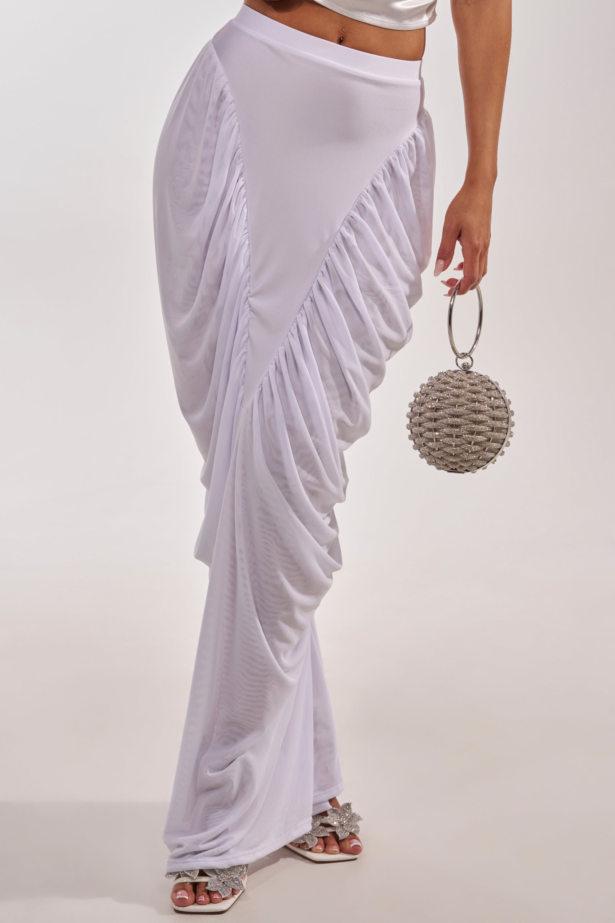 Wearing the DREAMY RUCHED SKIRT IN WHITE and silver sandals, a person holds a round, beaded handbag with a metal handle; only the lower half of their body is visible. 2