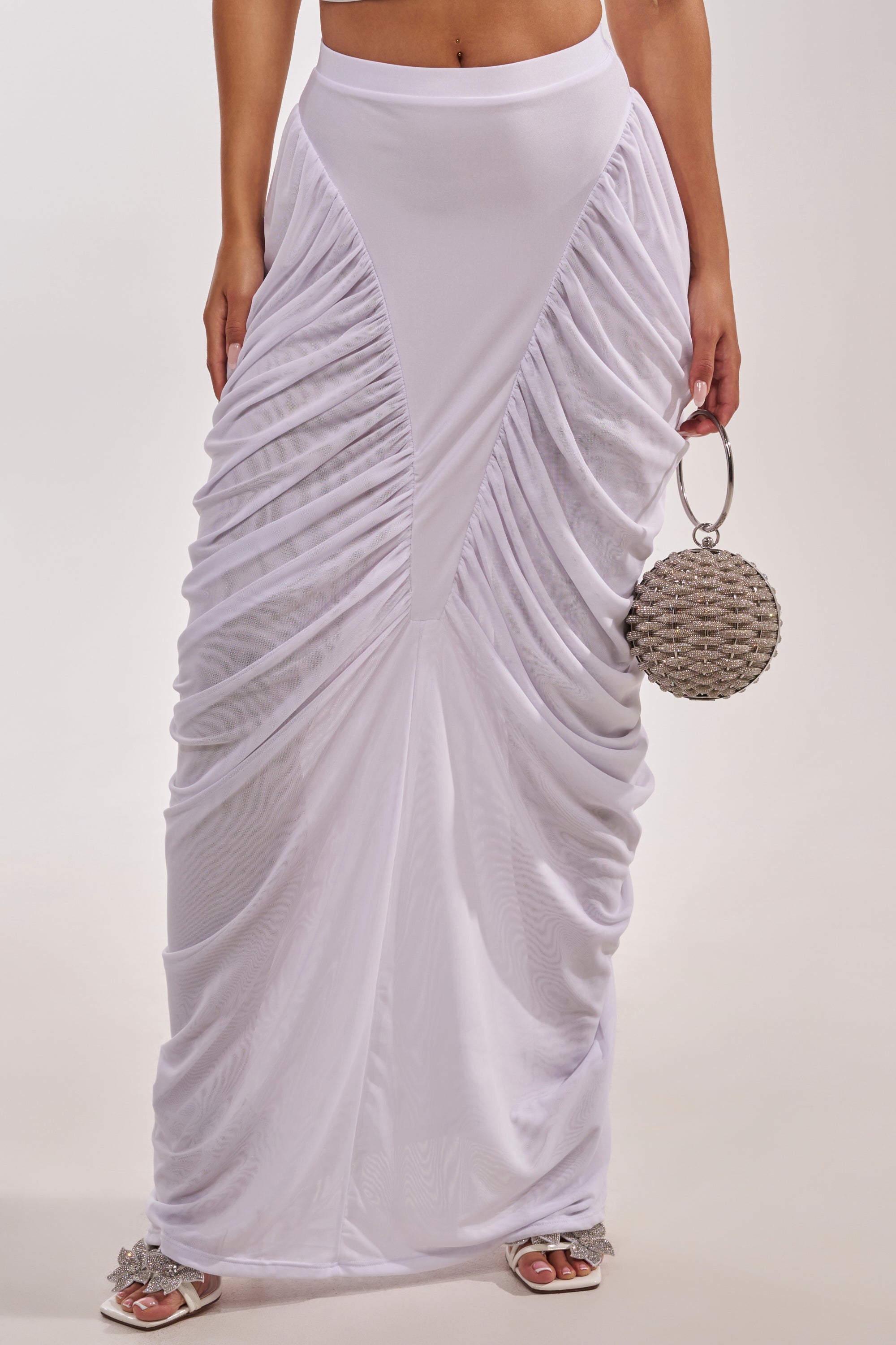 Wearing the DREAMY RUCHED SKIRT IN WHITE— a high-rise white maxi with side gathers and a smooth waistband—plus white sandals and carrying a round, textured silver handbag. The upper body is not shown. 3