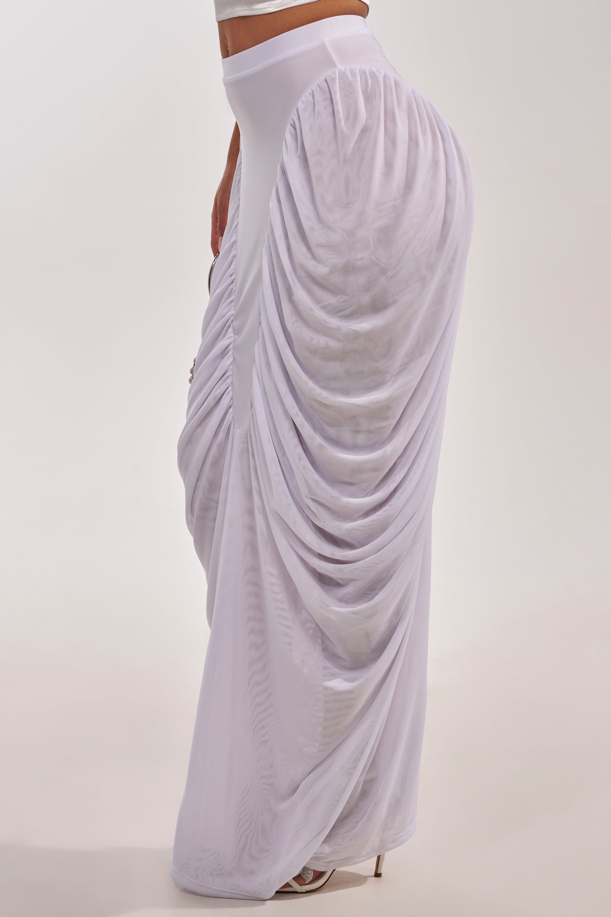 A person stands in profile against a plain background, wearing the DREAMY RUCHED SKIRT IN WHITE—a high-waisted maxi with sheer fabric and gathered side detailing. 4