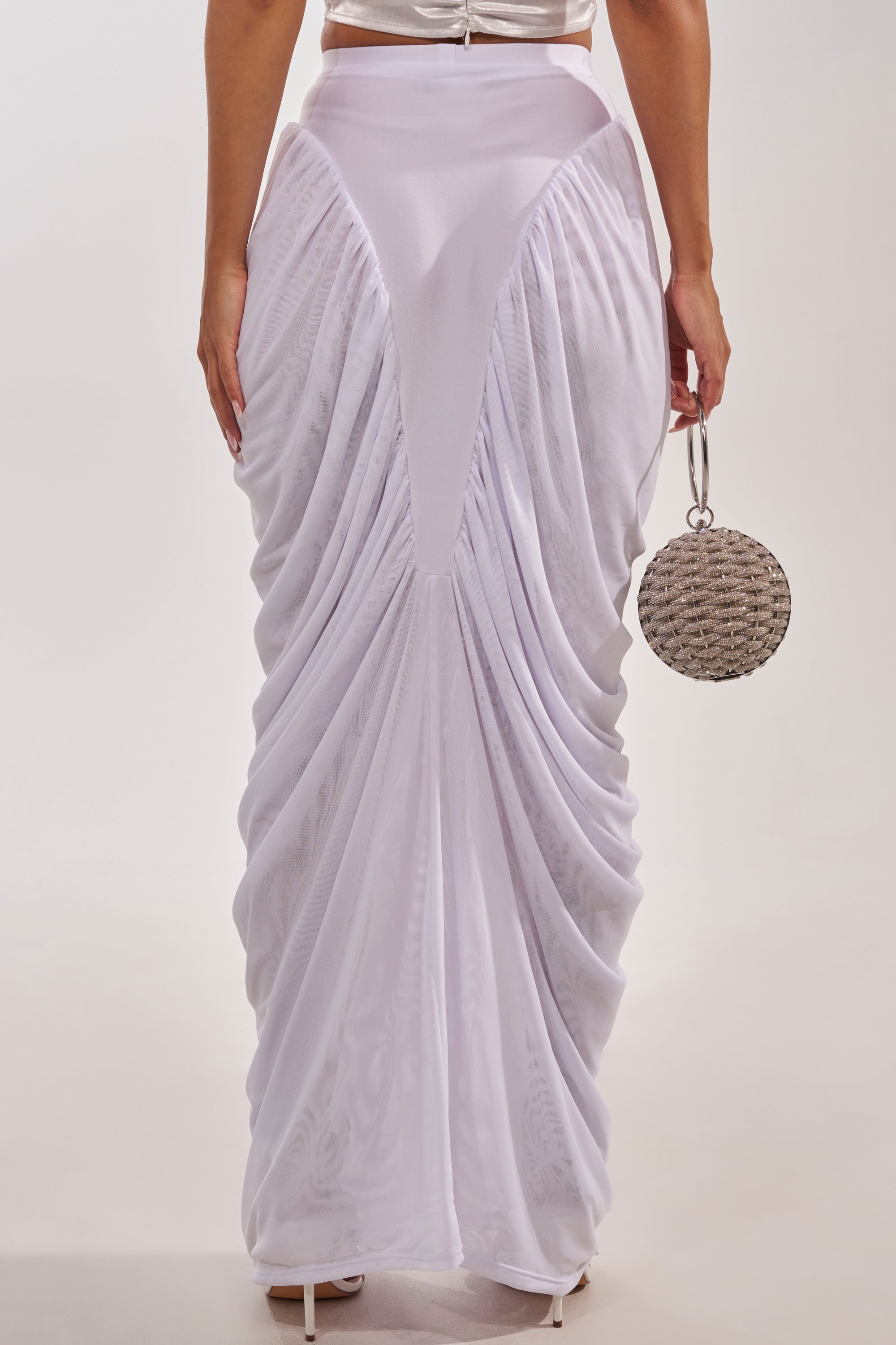 Wearing the DREAMY RUCHED SKIRT IN WHITE with gathered details, white heels, and holding a round woven handbag, the person is shown from the back against a plain background. 5