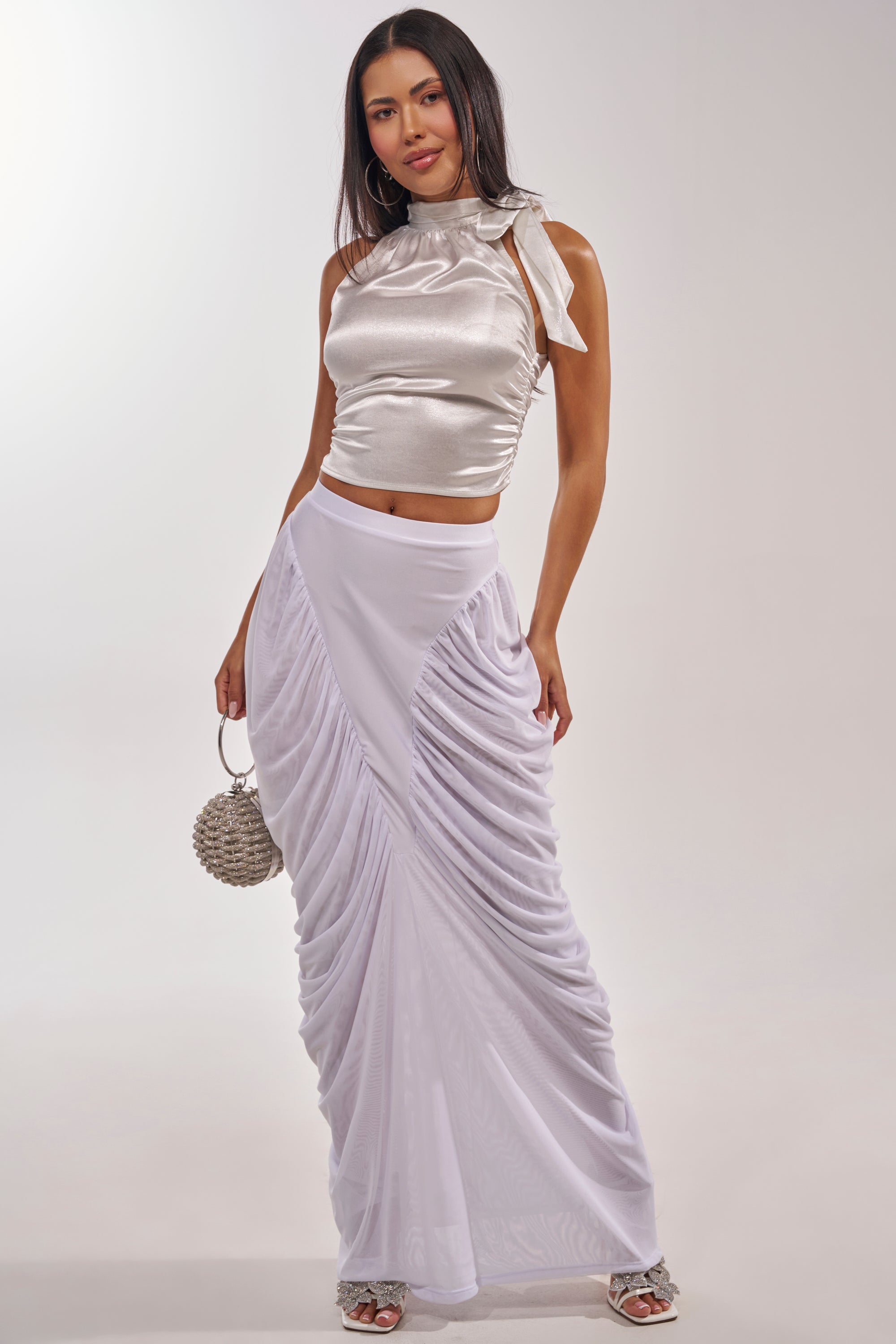 A woman wears the DREAMY RUCHED SKIRT IN WHITE with a shiny silver sleeveless crop top, holding a round beaded handbag and wearing open-toe heels, posing against a plain light background. 6