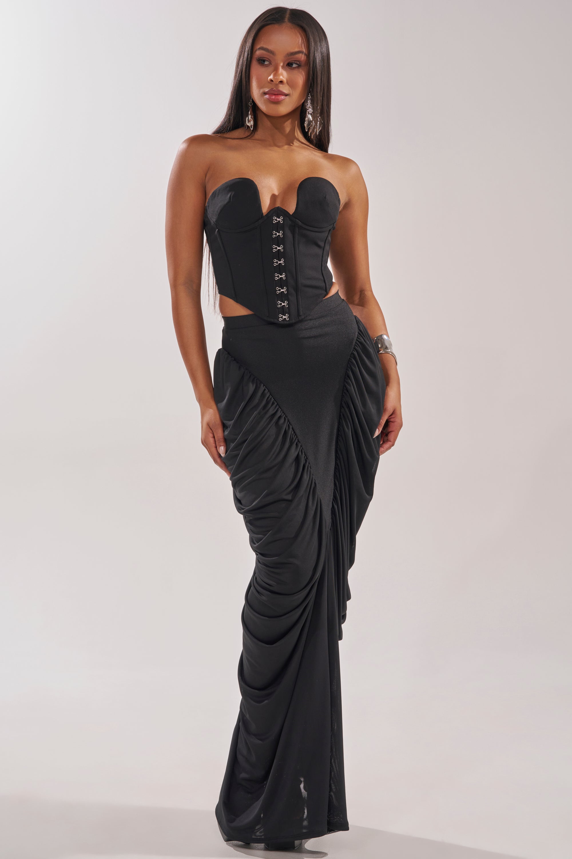 A woman models the DREAMY RUCHED SKIRT, a black strapless gown featuring a curve-hugging fit, corset-style bodice with front hooks, side cutouts, and a draped ruched skirt. She has long straight hair and accessorizes with earrings and a bracelet. 0
