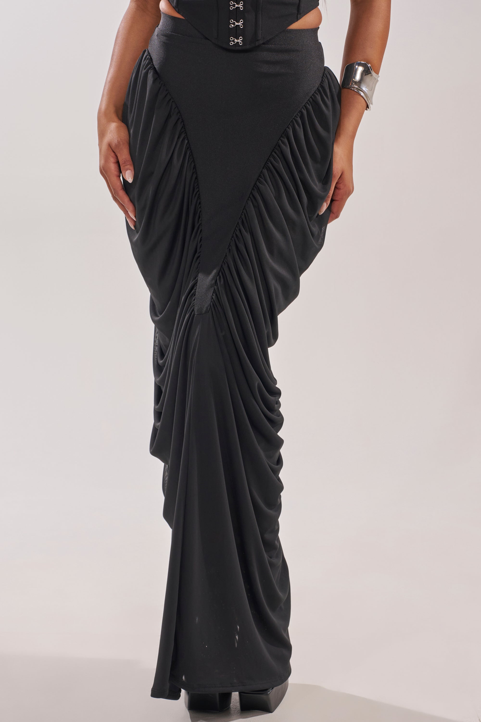 A person models the DREAMY RUCHED SKIRT—a dramatic black maxi with V-shaped draped fabric and wide waistband—styled with a silver cuff bracelet, highlighting the skirt and lower torso. 1