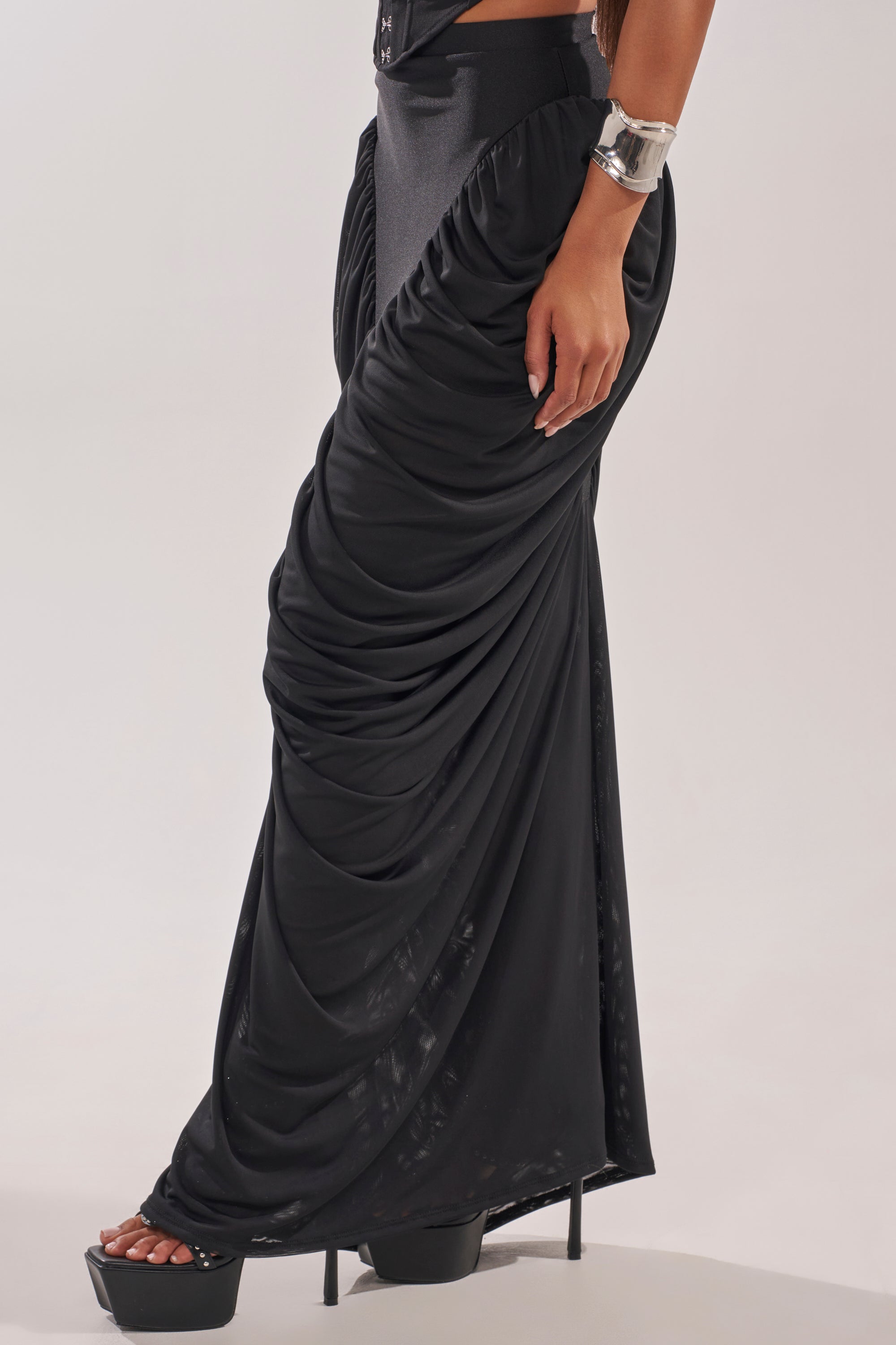 Someone wears the DREAMY RUCHED SKIRT with a high-rise waistband and black platform heels. A metallic cuff bracelet accents their wrist as they pose with a hand on their hip against a plain, light background. 3