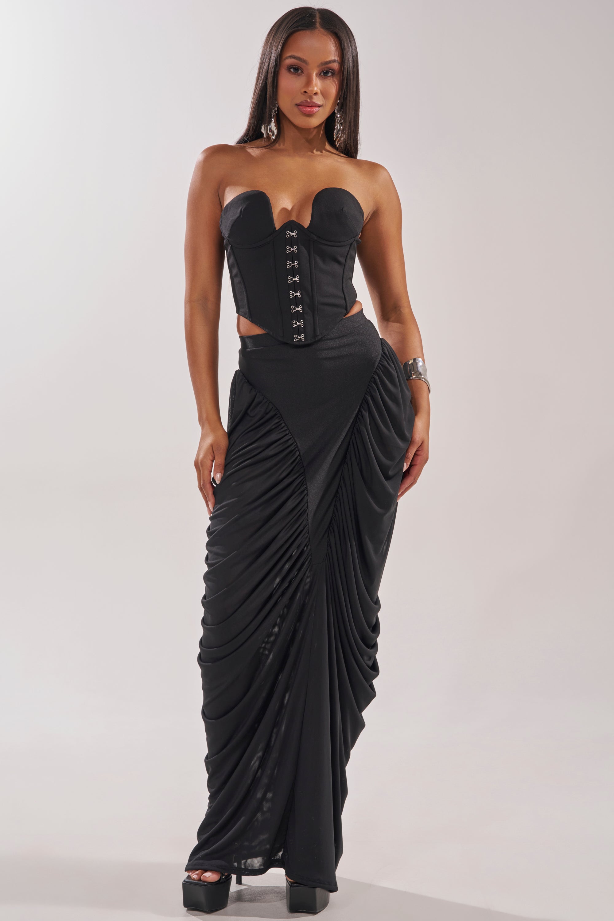 A woman wears a strapless black corset top with front clasps and the DREAMY RUCHED SKIRT. She has long straight hair, hoop earrings, a cuff bracelet, and black platform heels, standing against a plain background. 6