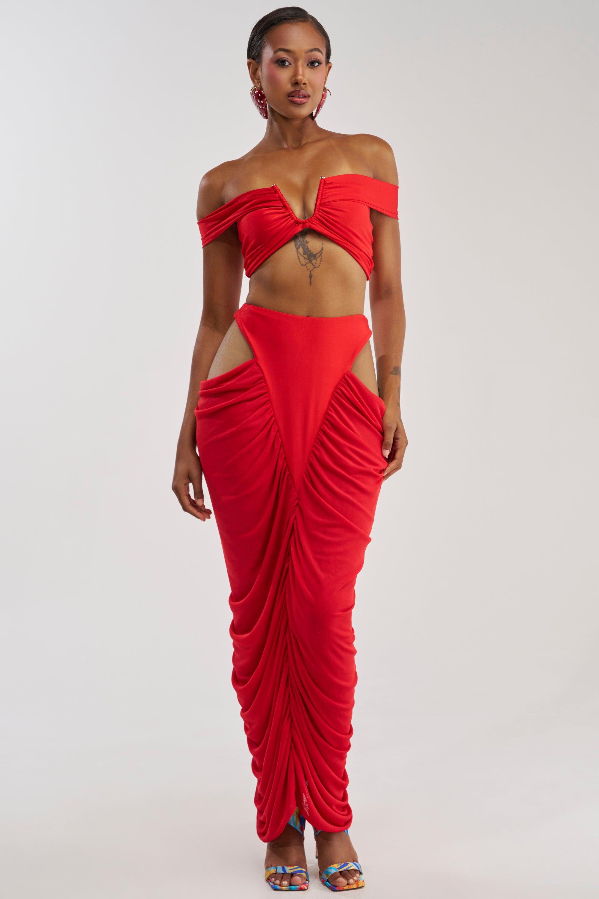A woman poses against a plain white background, wearing the HOTTIE ALERT MESH MAXI SKIRT IN RED. Styled with colorful sandals and statement jewelry, she shows off the skintight, ruched detailing and exposed hips of the vibrant red skirt, which features a flattering body-con mermaid fit that accentuates her silhouette.