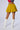 A person is facing away from the camera, wearing a yellow DONT LOOK BACK VELVET MINI SKIRT with a high-rise waist, white knee-high boots, and a cropped top featuring red, white, and blue sections. Tattoos are visible on the backs of their thighs. The background is plain and light-colored.